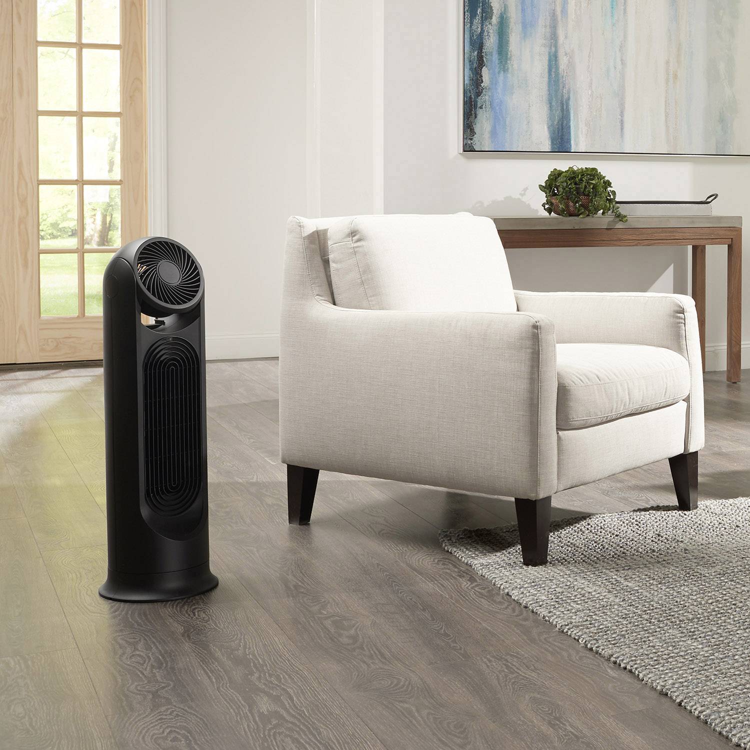 Černý sloupový ventilátor stojí v moderním obývacím pokoji vedle béžového křesla na dřevěné podlaze.