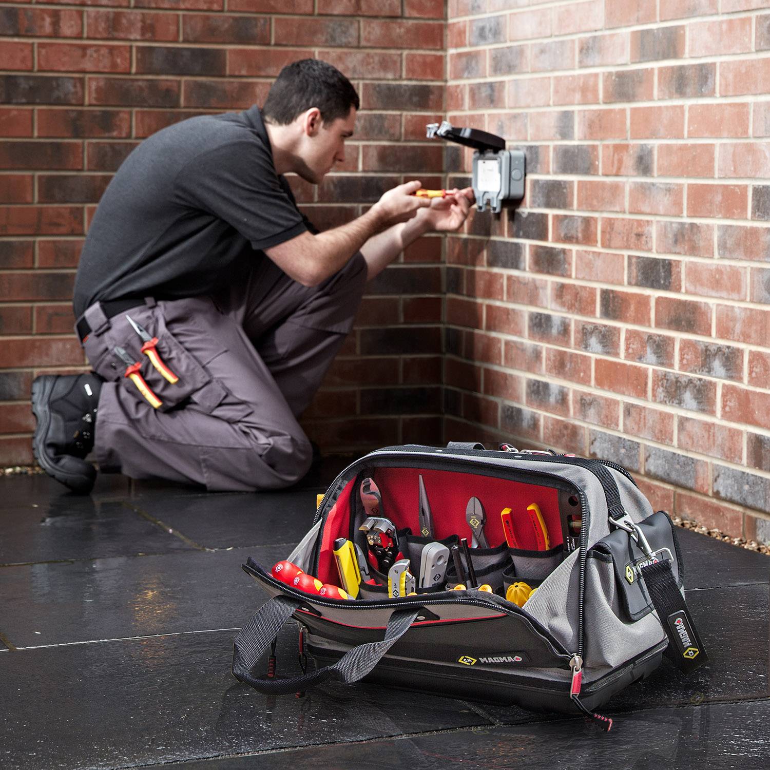 Elektrikář pracuje na zásuvce na cihlové zdi, s nástroji v otevřené brašně na nářadí v popředí.