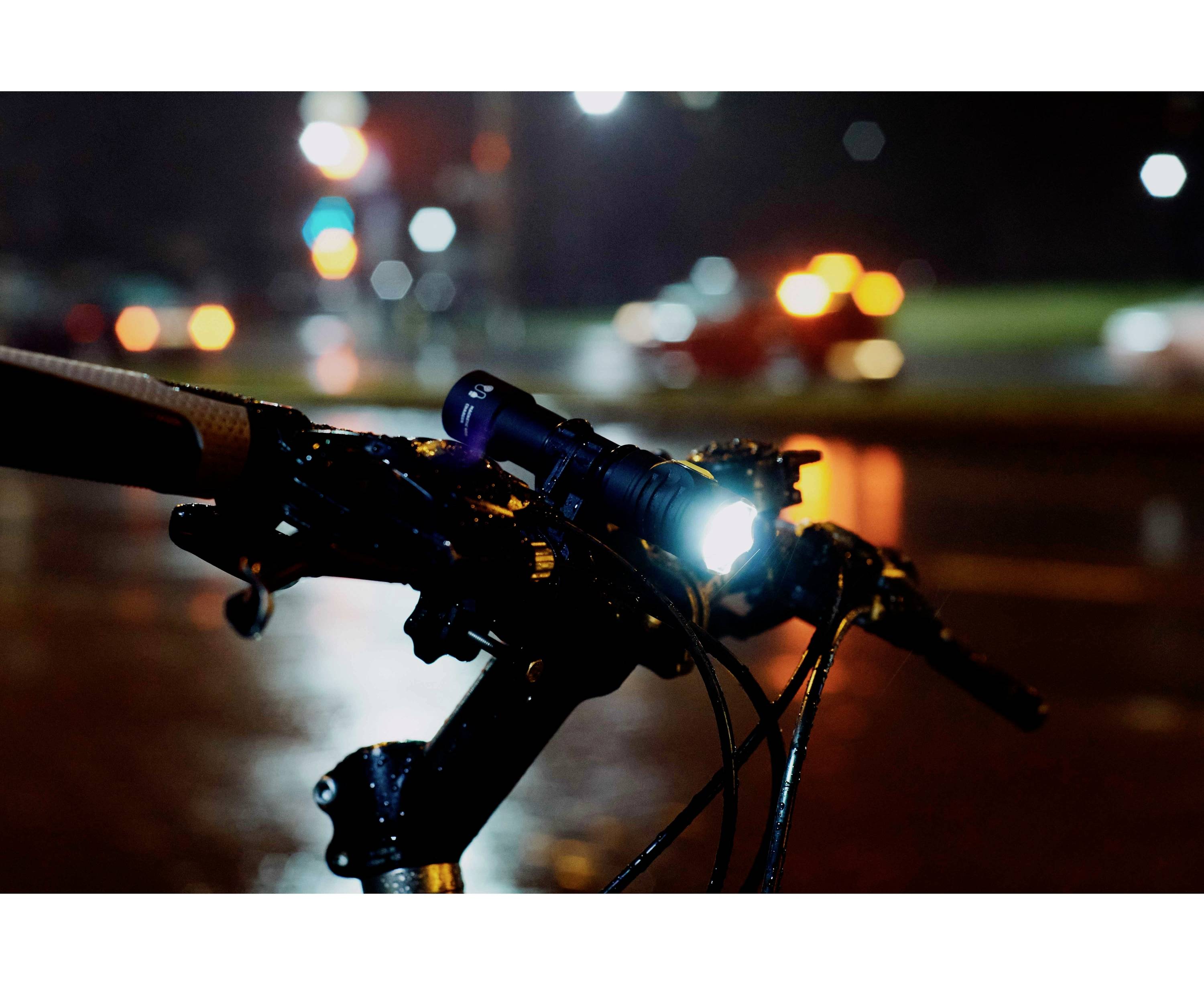 Řídítka jízdního kola se zapnutým světlem v noci za deště, rozmazaná světla projíždějících aut v pozadí.