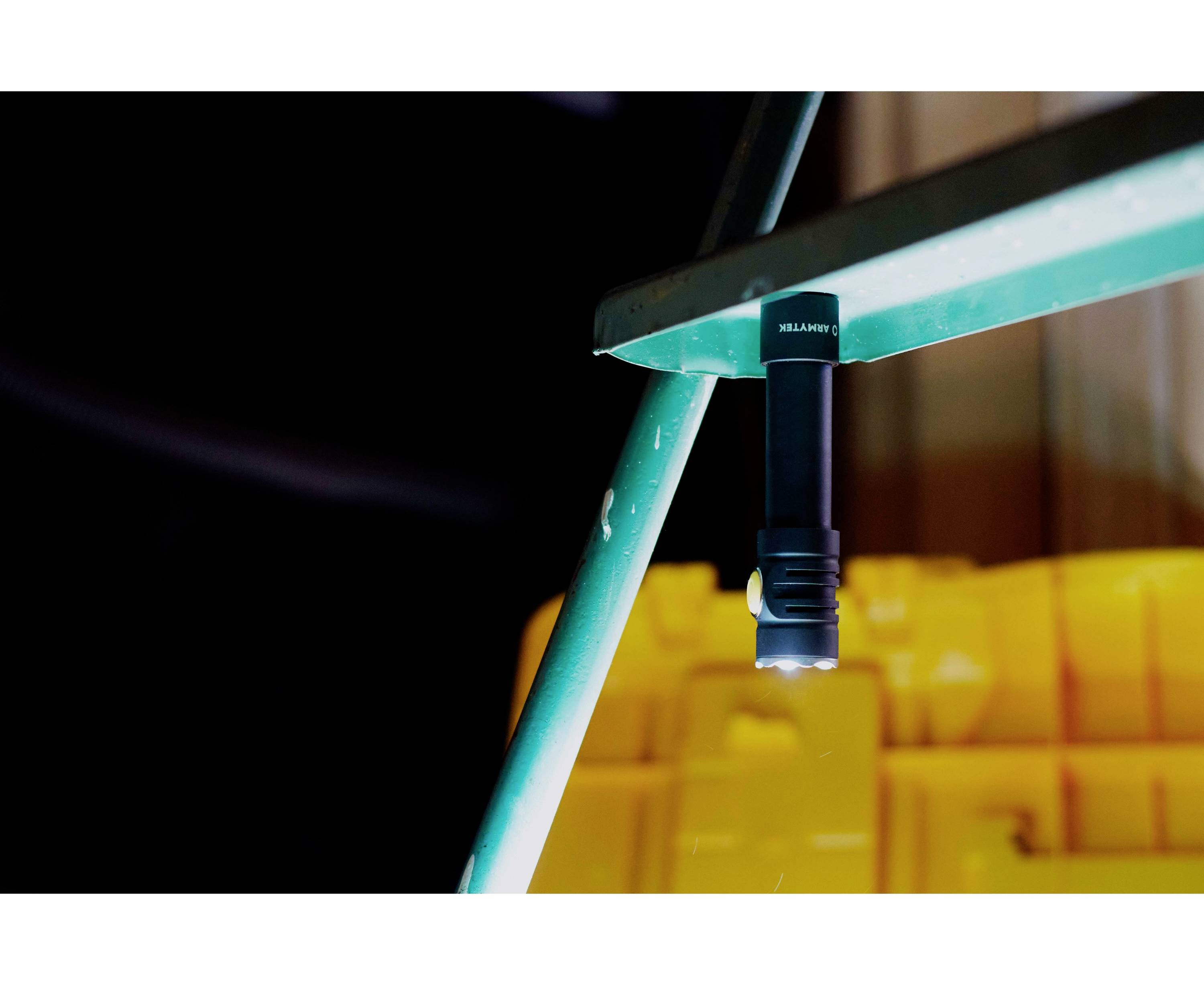 Černá svítilna je magneticky připevněná k zelenému kovovému regálu, pozadí je rozostřené s žlutým kontejnerem.