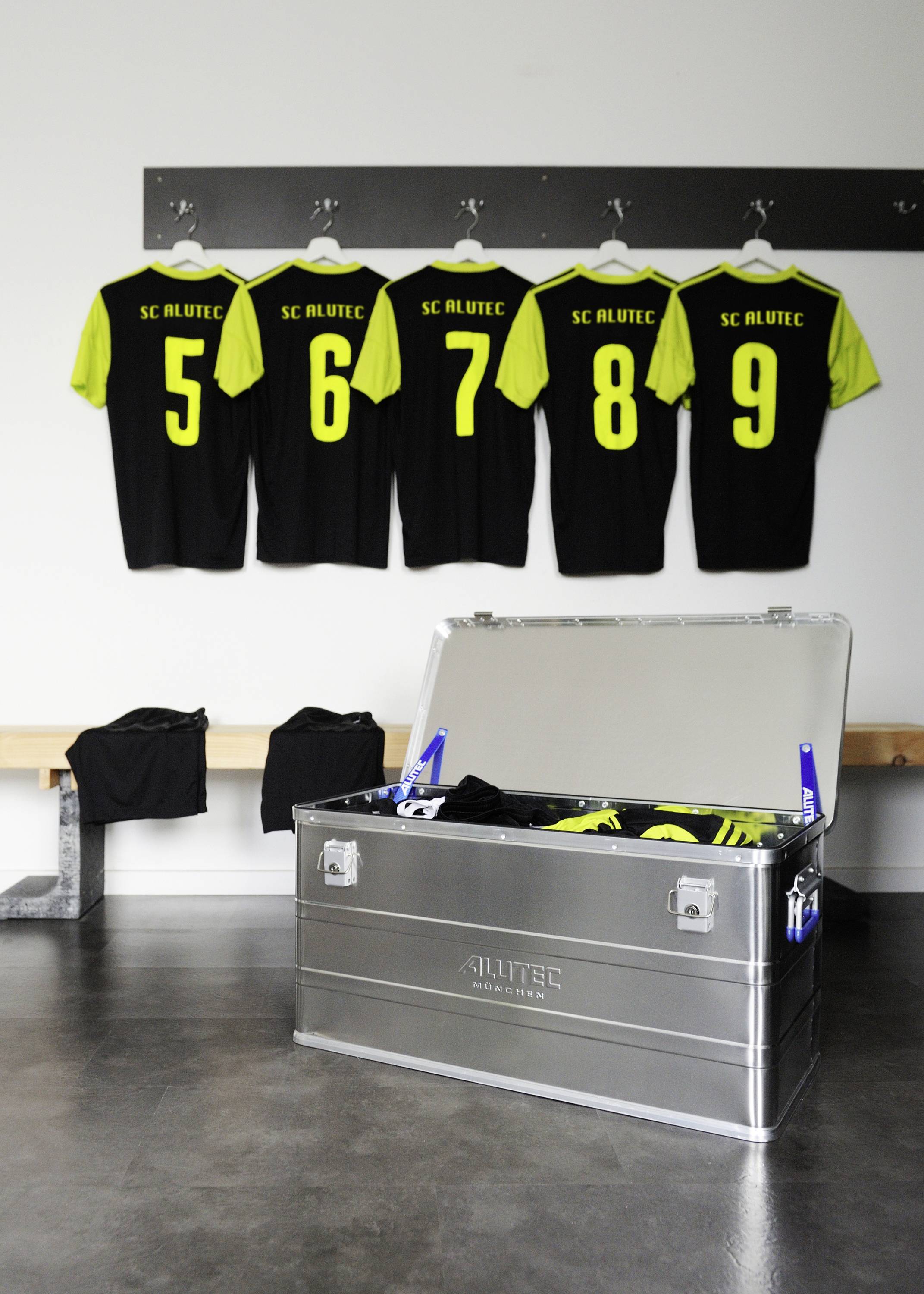 Pět dresů s čísly 5 až 9 visí v šatně. Pod nimi stojí kovový kontejner s dalšími sportovními potřebami.