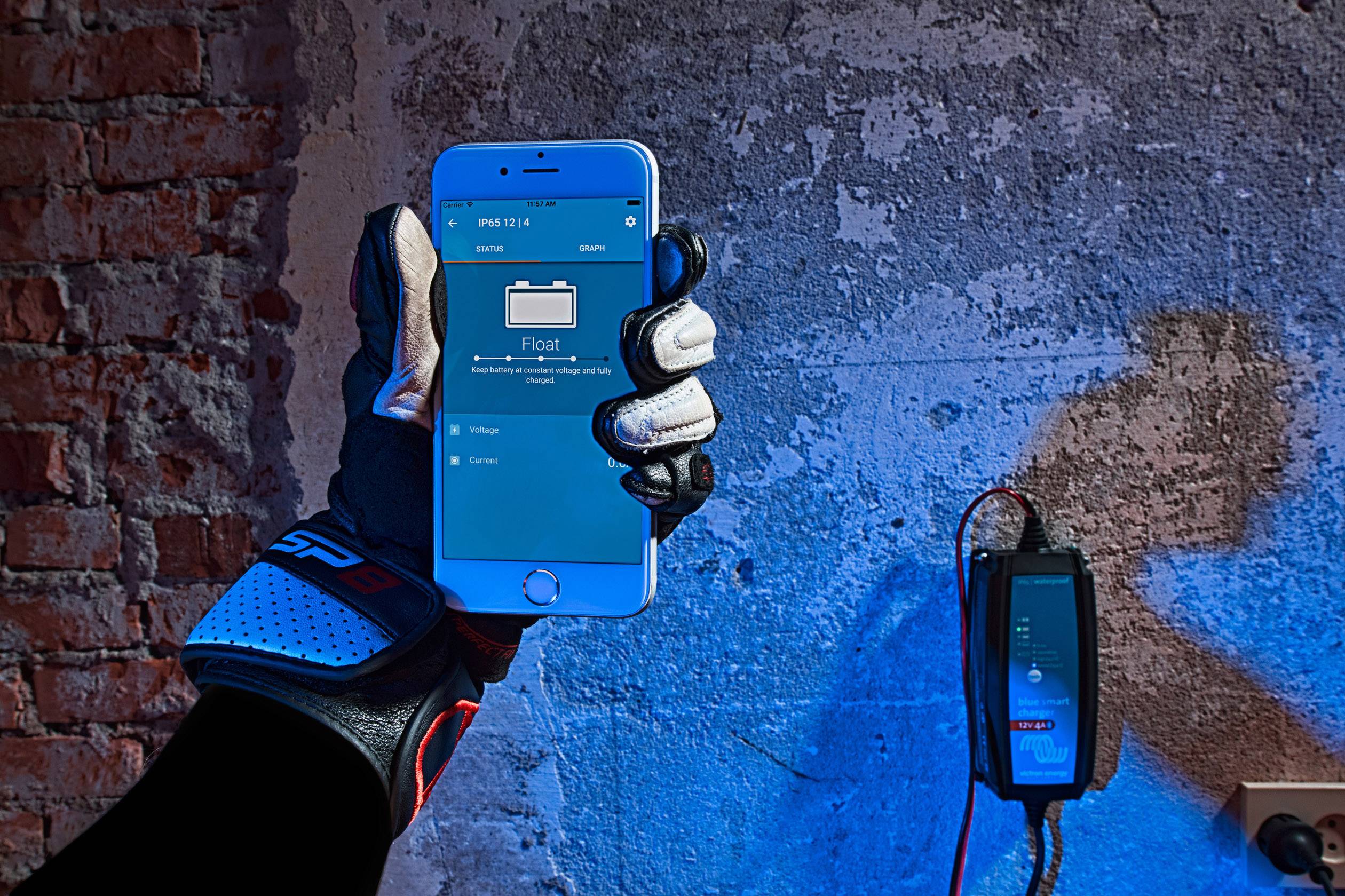 Rukavicí opatěná ruka drží smartphone s otevřenou aplikací před osvětlenou stěnou. Vpravo visí elektronické zařízení na zdi.