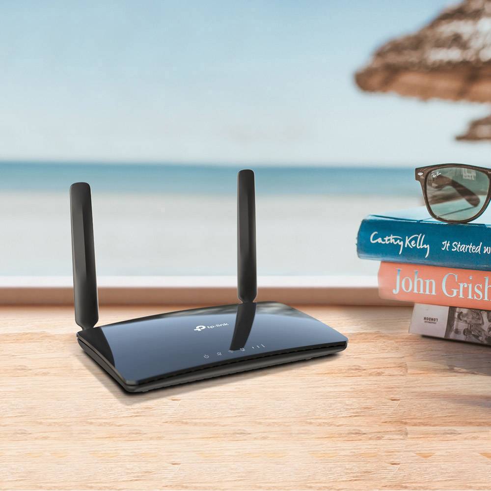 Router Wi-Fi se dvěma anténami stojí na stole vedle hromady knih. V rozmazaném pozadí je vidět moře a obloha.