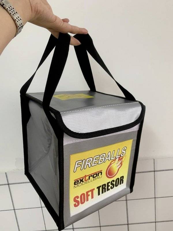 Ruka drží šedou tašku s držadly, na které je napsáno 'Fireballs' a 'Soft Tresor'. Taška je lehce nadzvednutá.