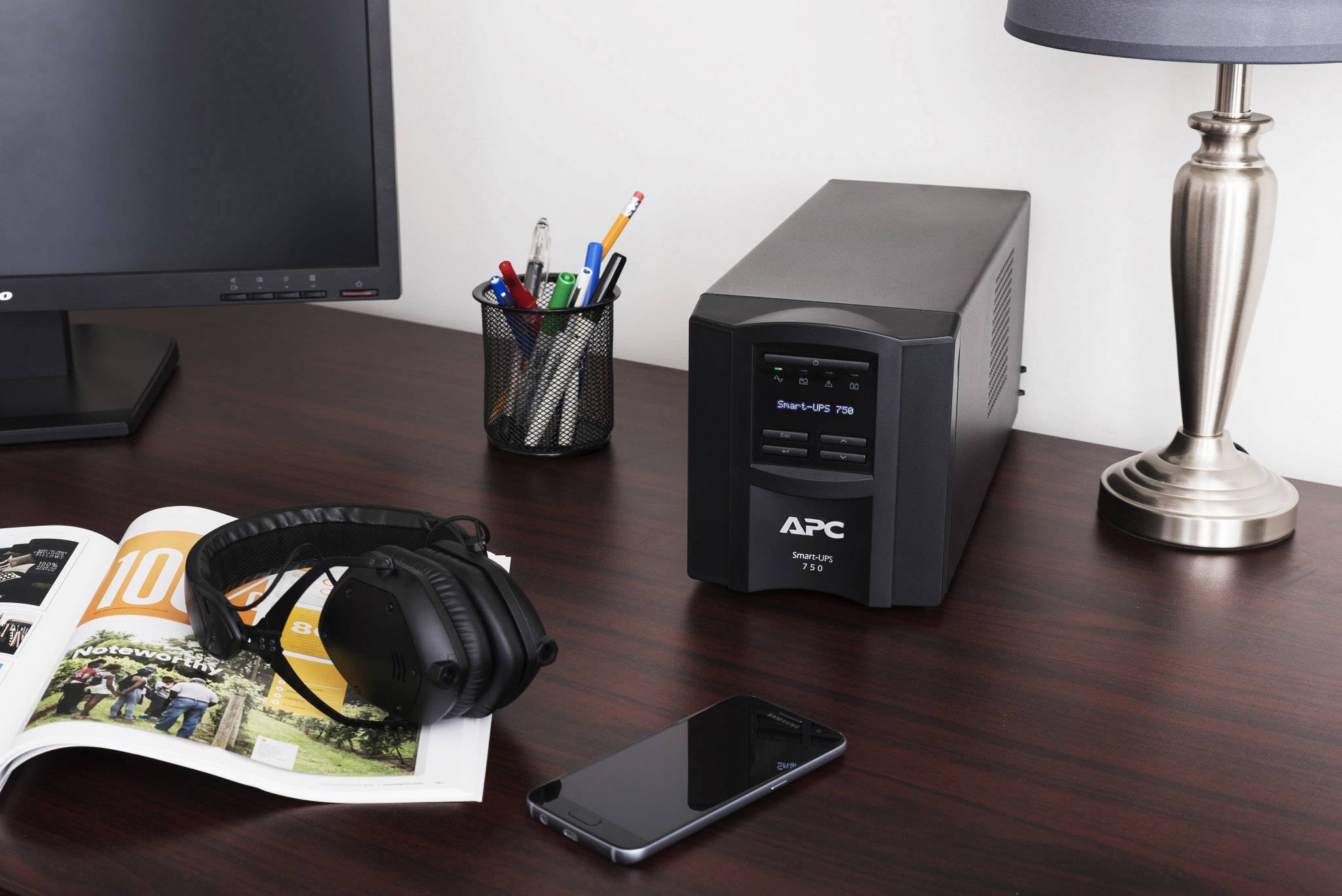 Psací stůl s otevřeným časopisem, sluchátky, smartphonem, lampou, monitorem a záložním zdrojem napájení (APC) vedle stojánku na tužky.