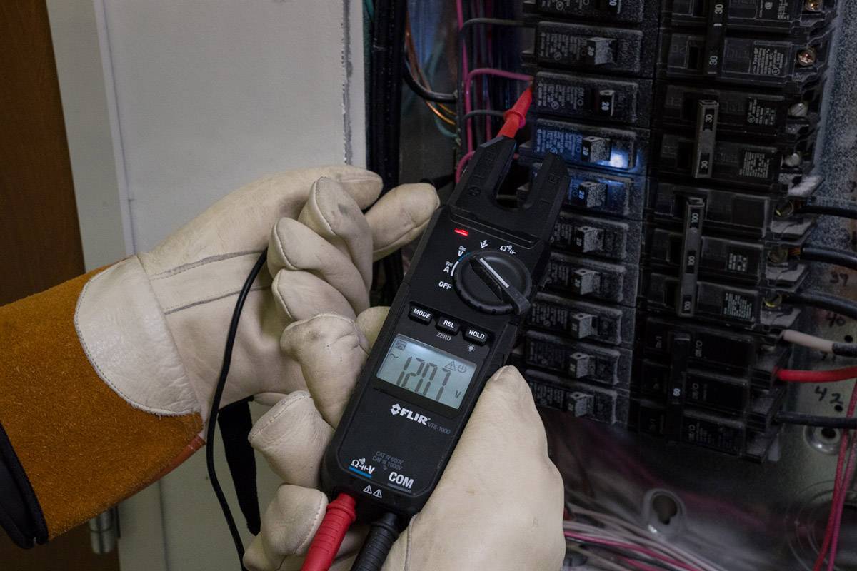 Osoba měří elektrické napětí multimetrem na rozvodné skříni. Displej ukazuje '120V'.