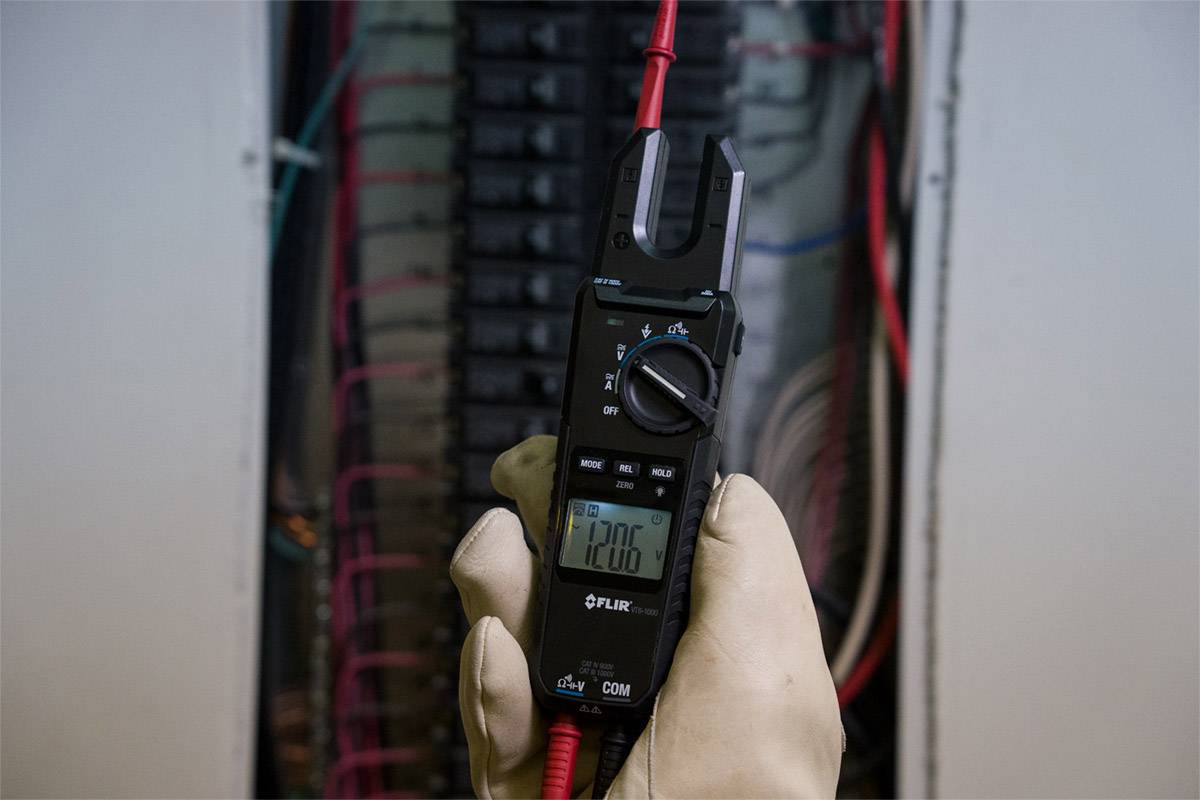 Osoba drží multimetr u elektrické rozvodné skříně a měří 120,6 voltů. Používají se červené a černé kabely.