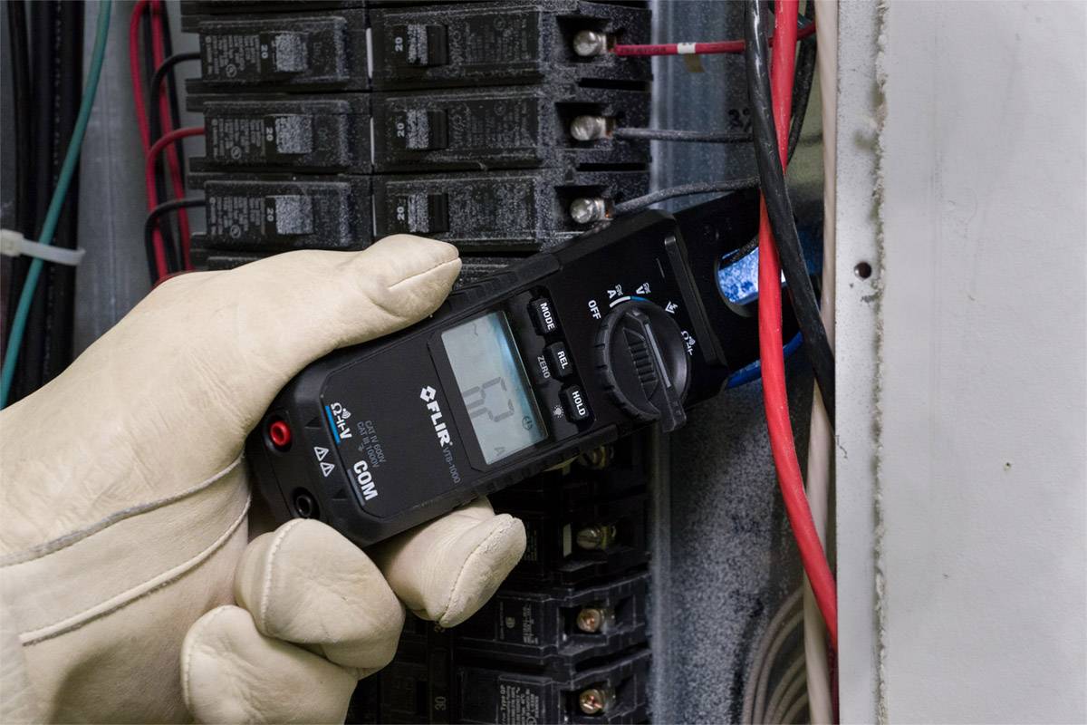 Osoba v ochranných rukavicích měří pomocí digitálního multimetru průtok elektrického proudu na elektrickém rozvaděči.