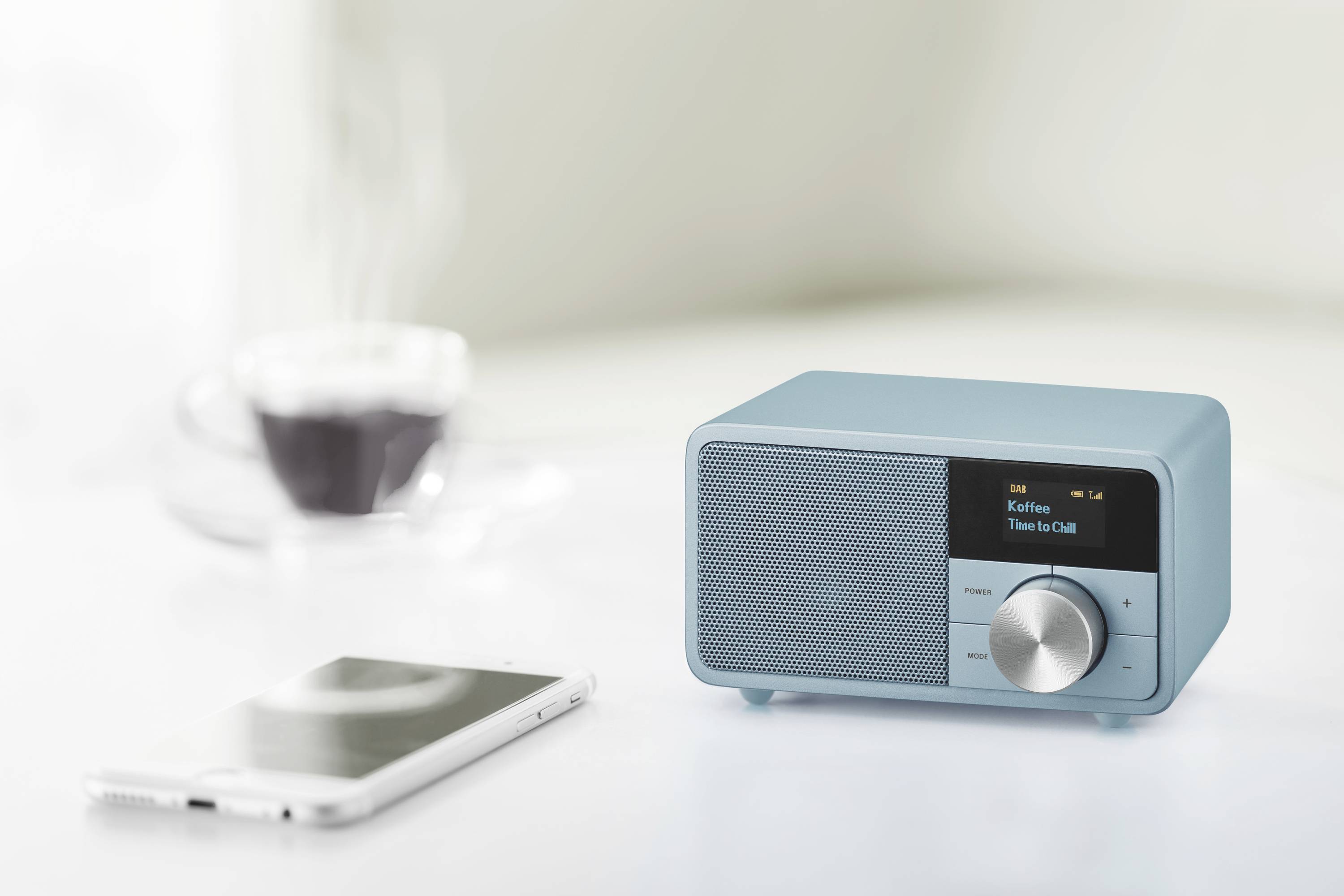 Modrý stolní radiopřijímač s displejem zobrazuje 'Interpret: Time to Chill'. Vpravo ovladač hlasitosti. V pozadí šálek a smartphone.