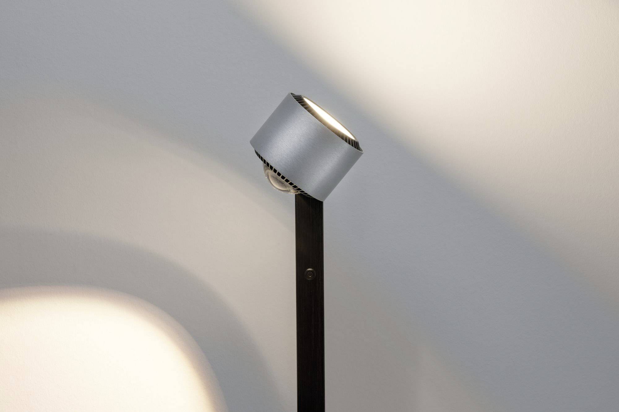 Moderní stříbrná stojací lampa vysílá teplé světlo na béžovou stěnu a vytváří měkké stíny.