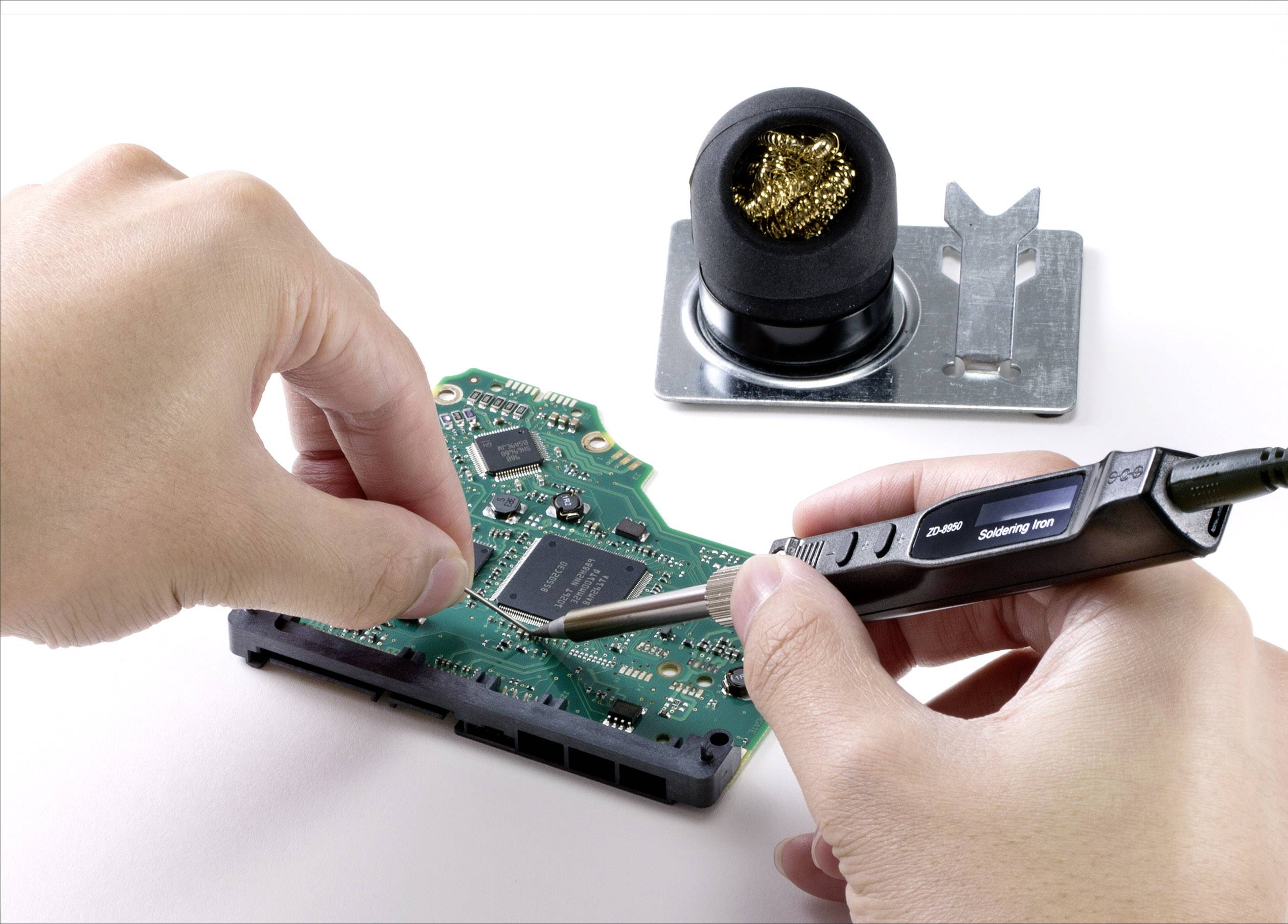 Osoba pájí komponenty pájecí pákou na zelené desce plošného spoje. V pozadí stojí držák pájecí houby.