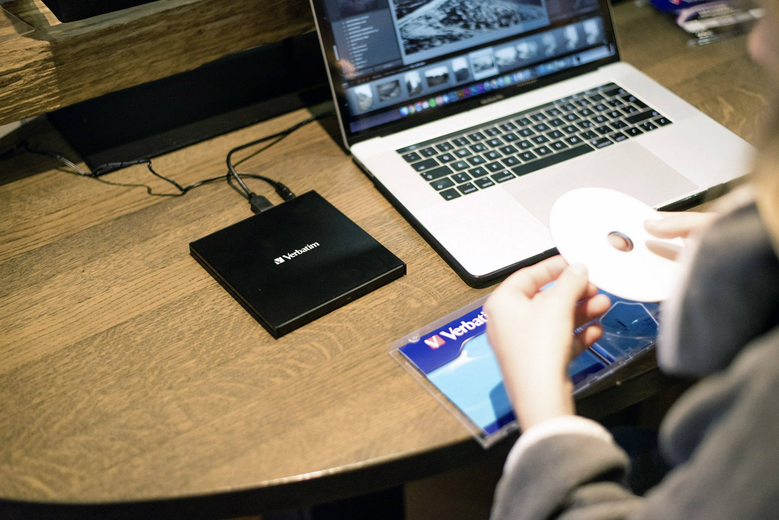 Osoba vkládá CD do mechaniky Verbatim na stolku s laptopem, který zobrazuje software pro úpravu obrázků.