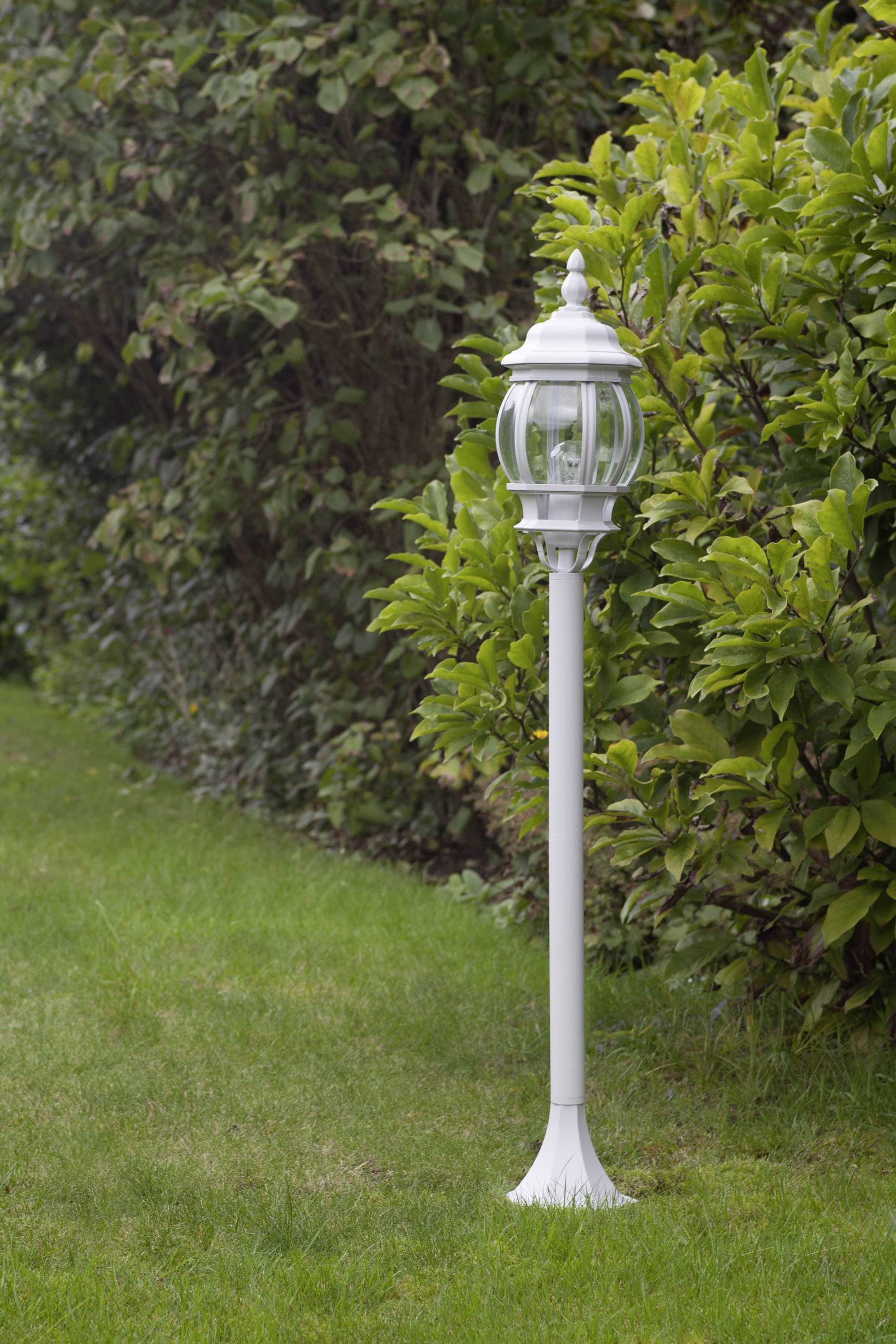 Bílá zahradní lucerna stojí na udržovaném trávníku vedle hustých zelených keřů a osvětluje cestu v zahradě.