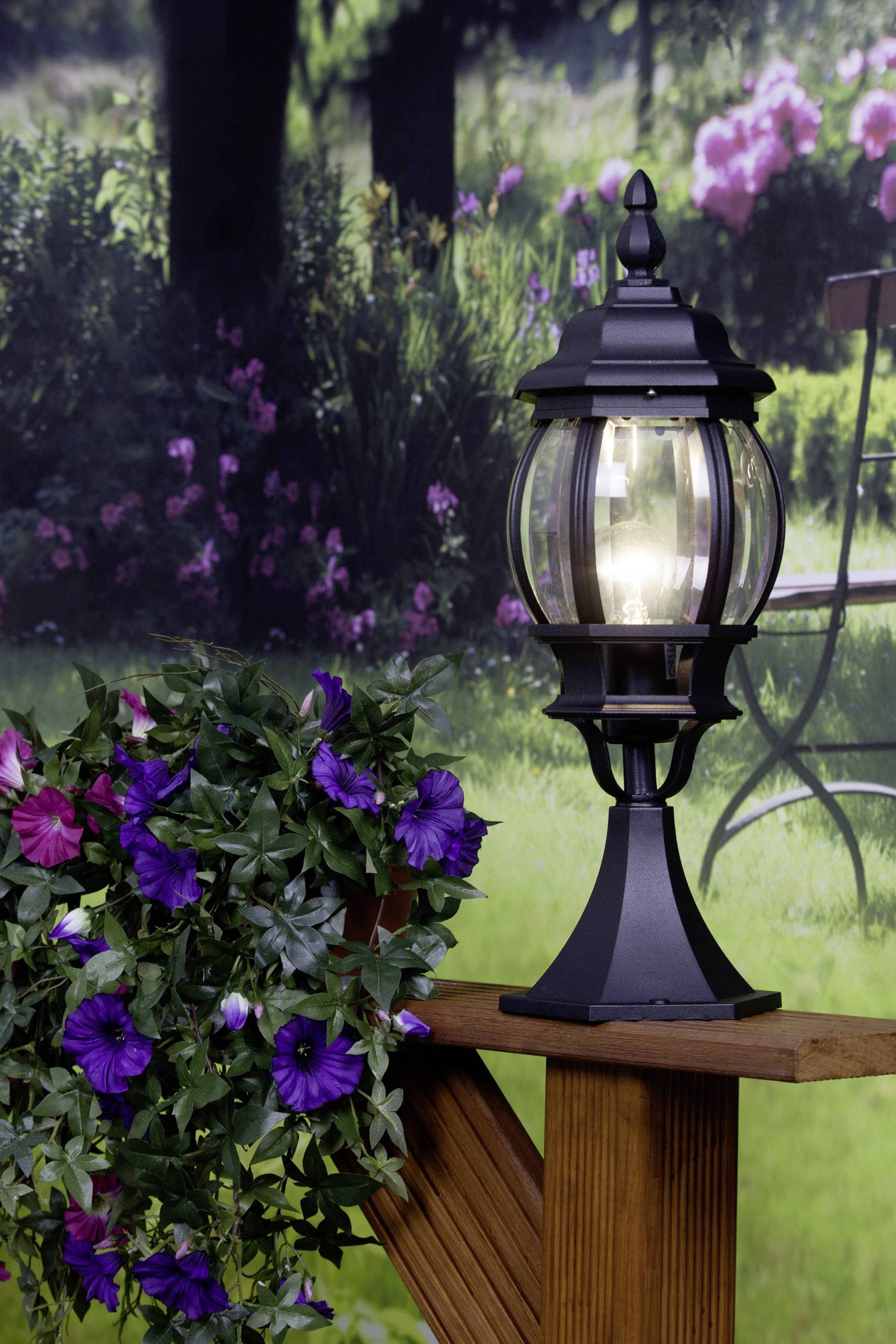 Černá lucerna svítí na dřevěném trámu vedle kvetoucího koberce z fialových květů v zelené zahradě.