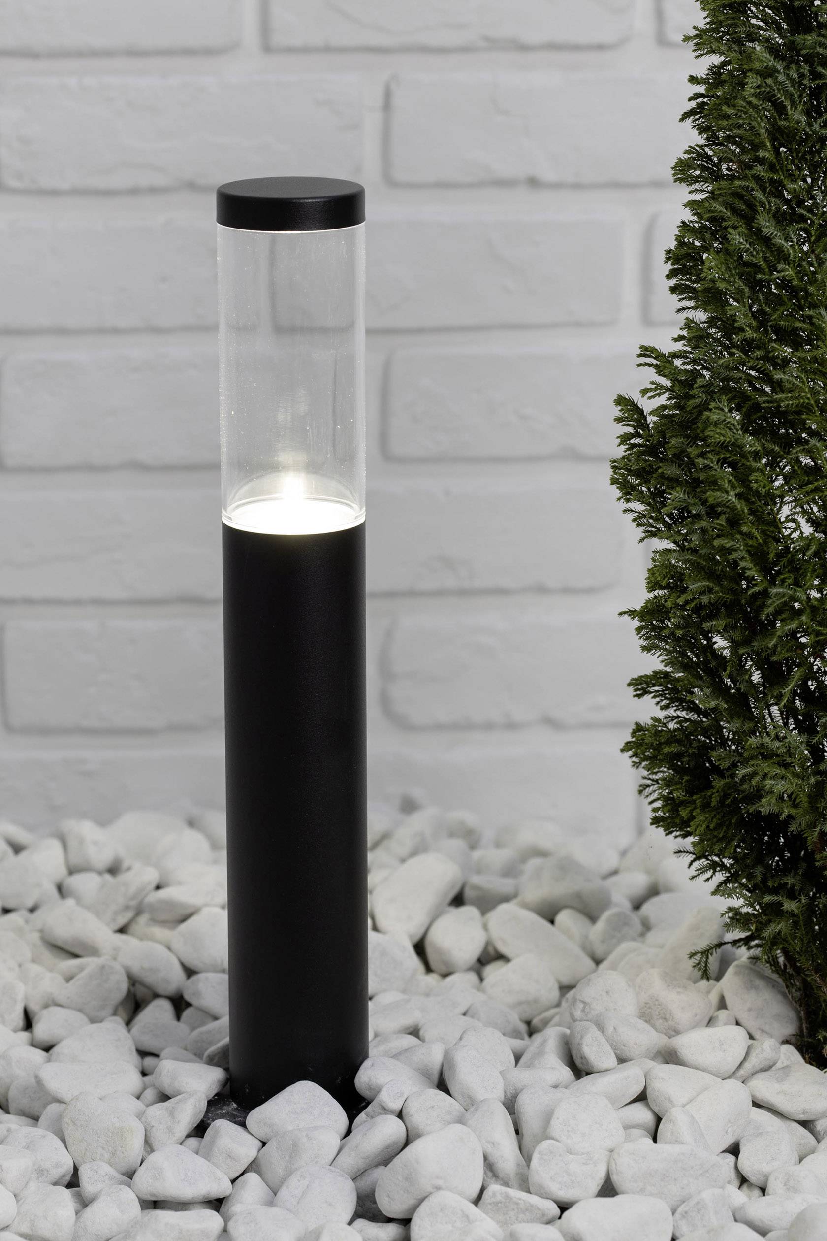 Černé zahradní světlo zapuštěné v oblázkovém štěrku vyzařuje jemné světlo. Vedle něj malý zelený keř před bílou cihlovou zdí.