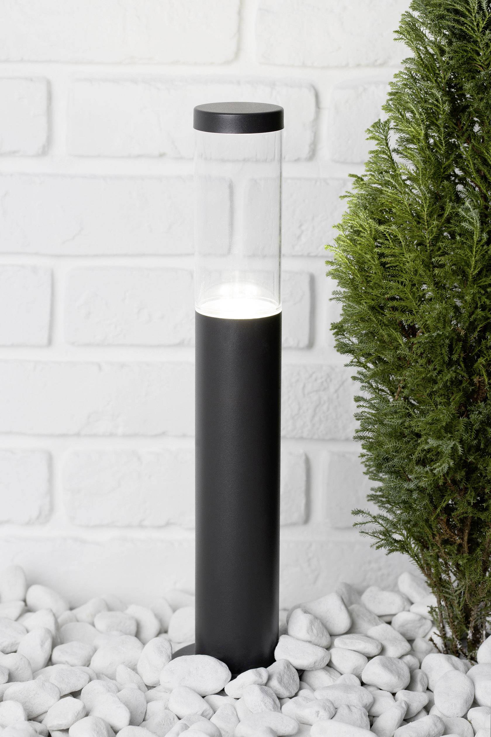 Moderní černá stojací lampa stojí na bílých oblázích vedle zeleného keře před bílou cihlovou zdí.