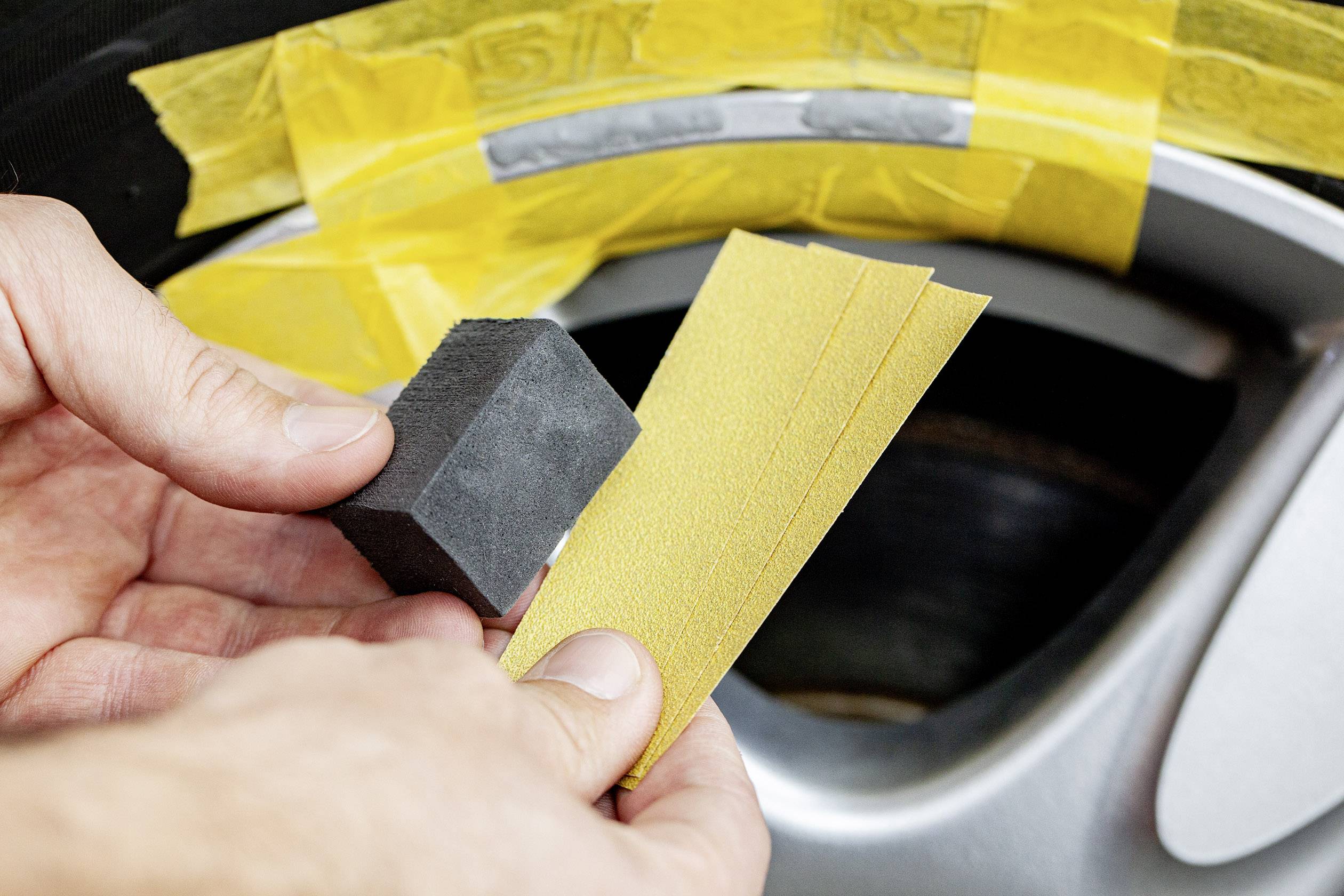 Detailní záběr rukou držících brusný nástroj a smirkový papír před pneumatikou automobilu zakrytou lepicí páskou, připravených k opracování.