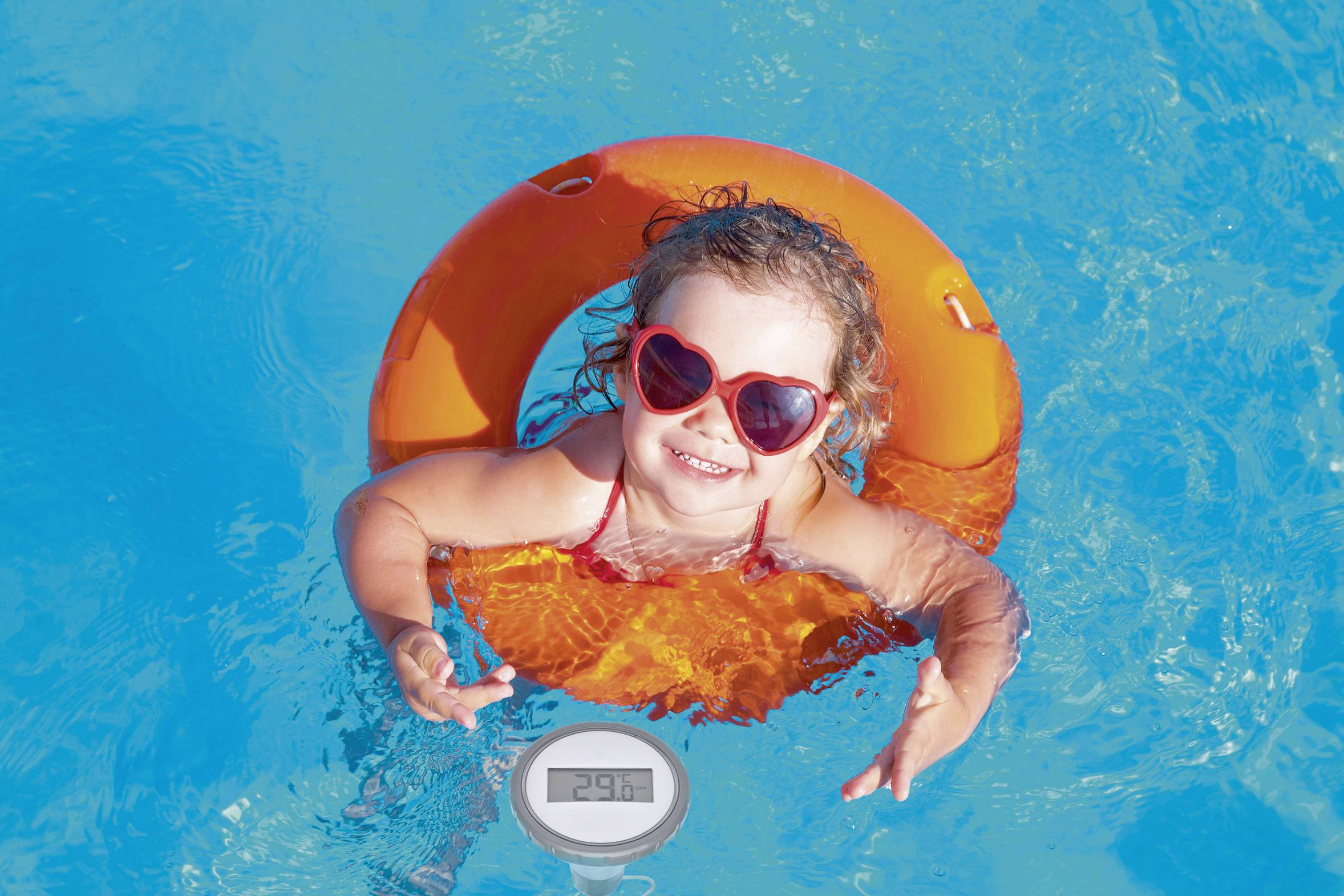 Malá usmívající se dívka v srdíčkových slunečních brýlích plave v modrém bazénu na oranžovém nafukovacím kruhu.