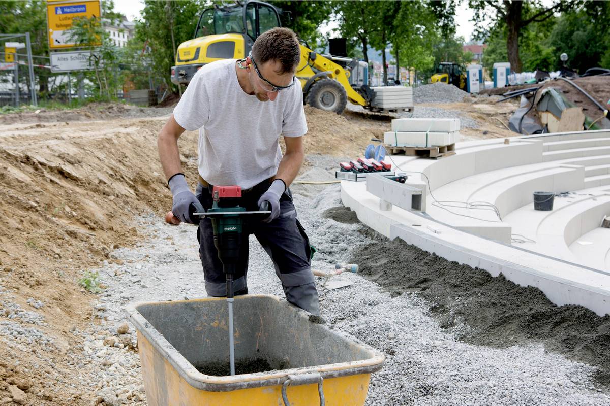 Dělník míchá beton ve vedru na staveništi. V pozadí jsou viditelné stavební stroje a stavební materiály.