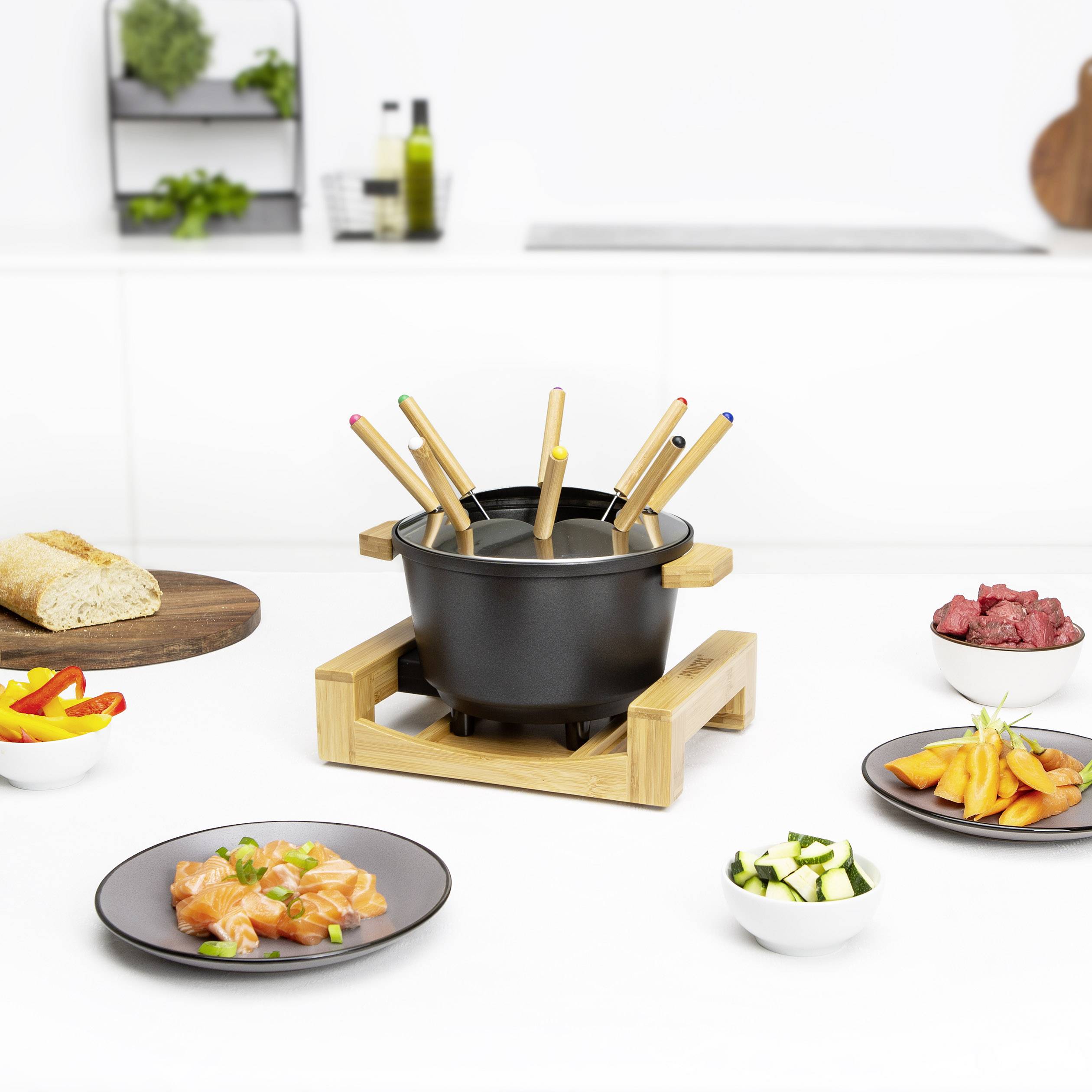Fondué set s dřevěným rámem uprostřed, obklopený talíři s chlebem, zeleninou a masem, na bílém kuchyňském stole.