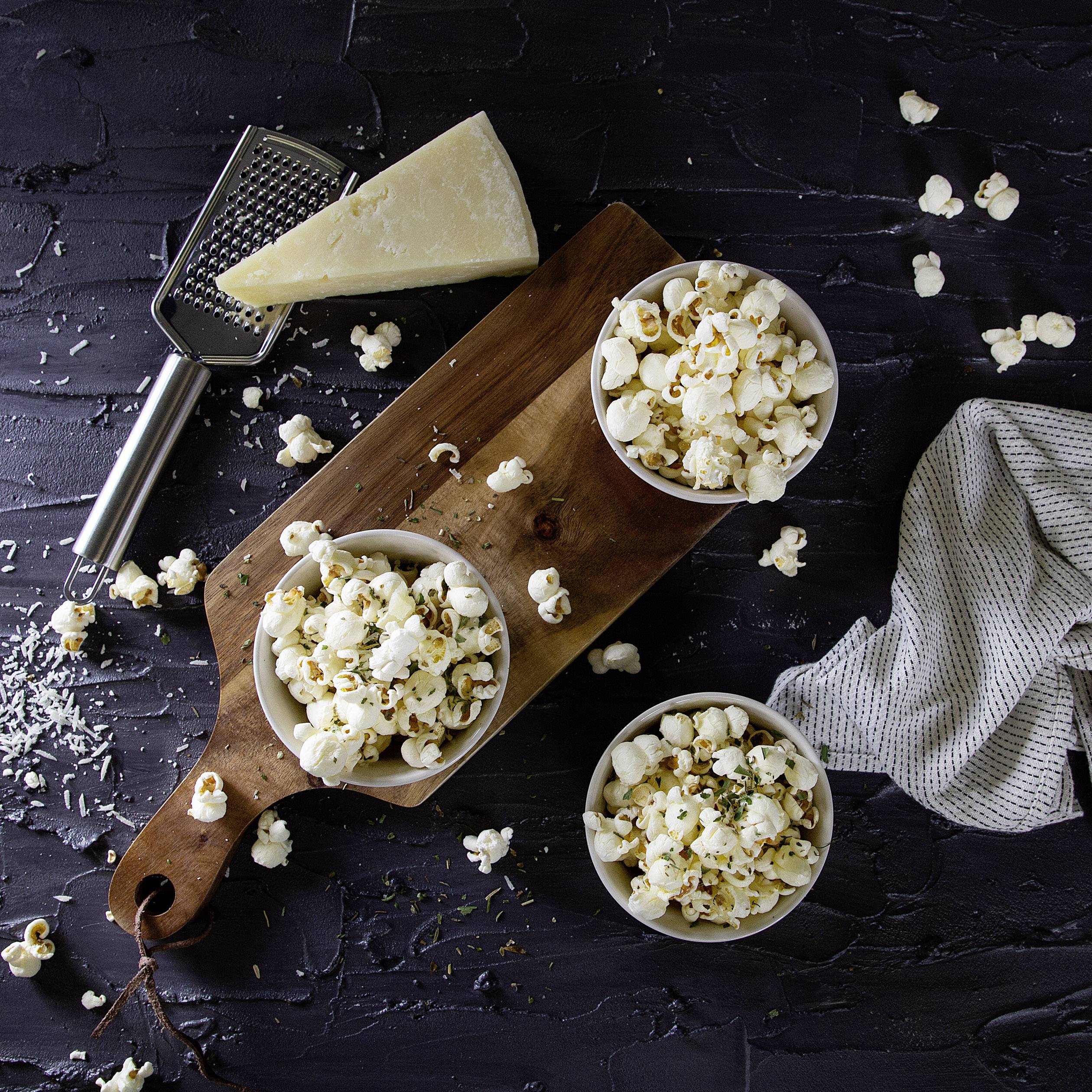 Tři misky s popcornem na dřevěném prkénku, obklopené nastrouhaným sýrem, struhadlem na sýr a kuchyňskou utěrkou.