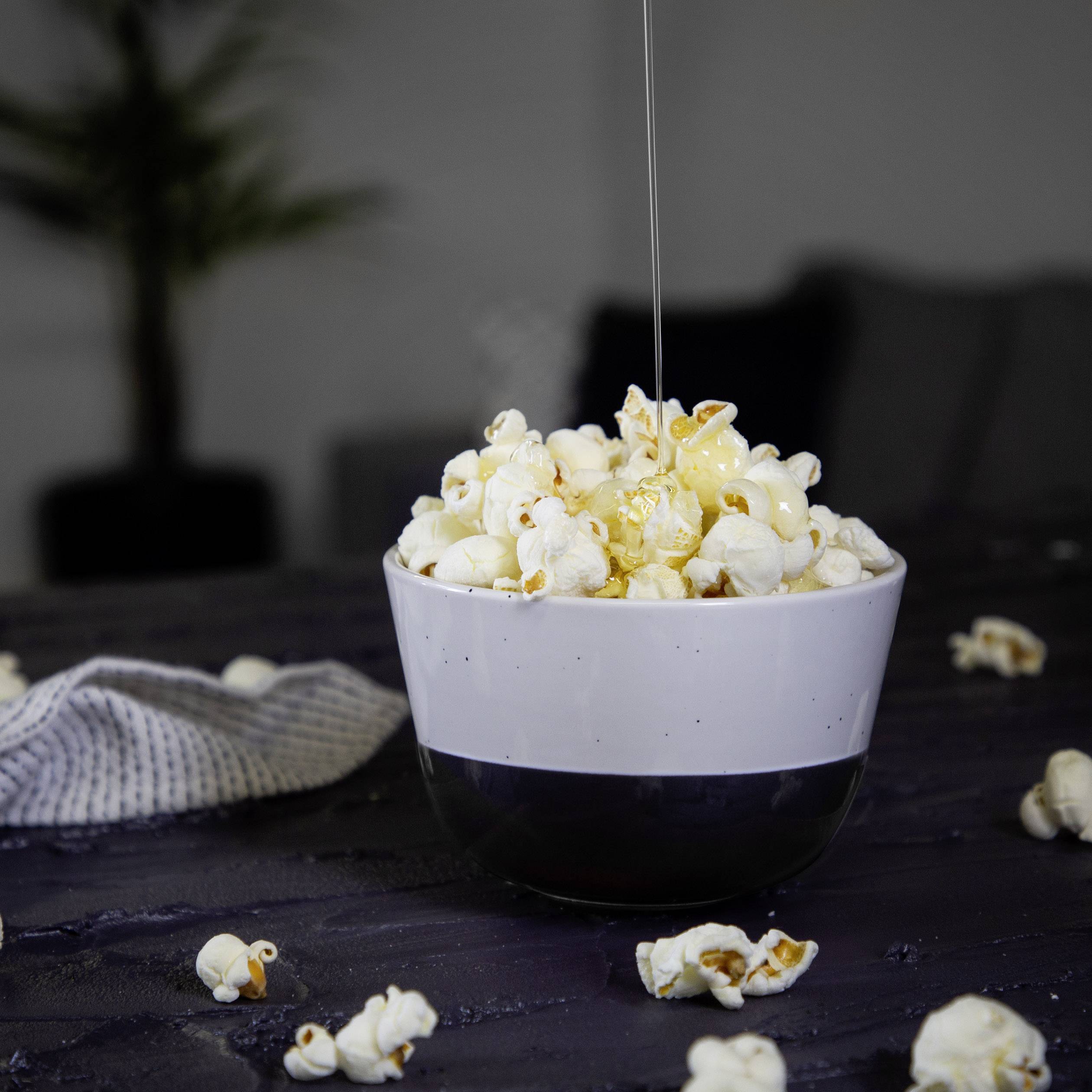 Miska s popcornem na stole, na kterou je nalévána medová poleva, s rozostřeným pozadím.