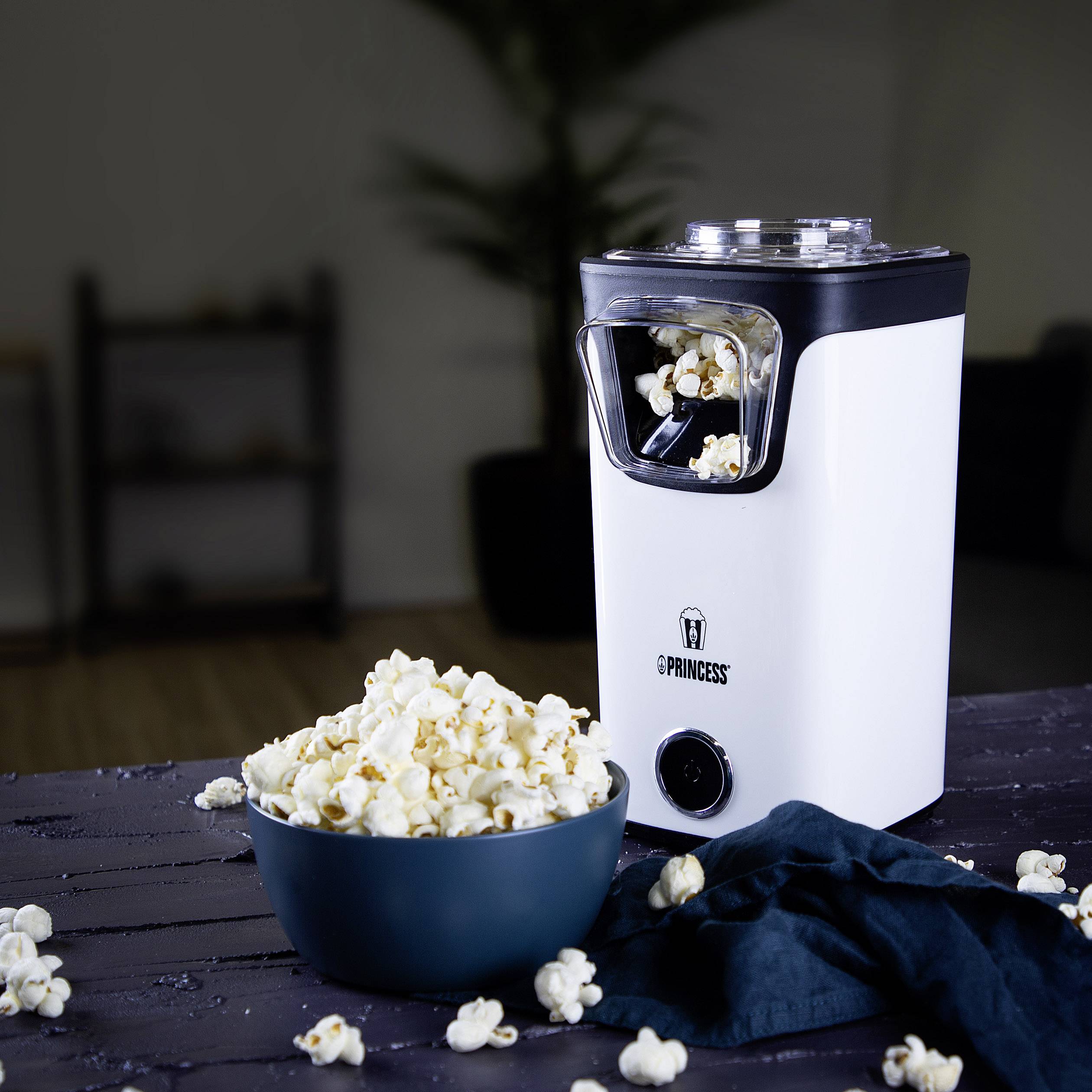 Bílý stroj na popcorn s mísou plnou čerstvého popcornu vedle něj. V pozadí jsou rozmazané police a rostliny.