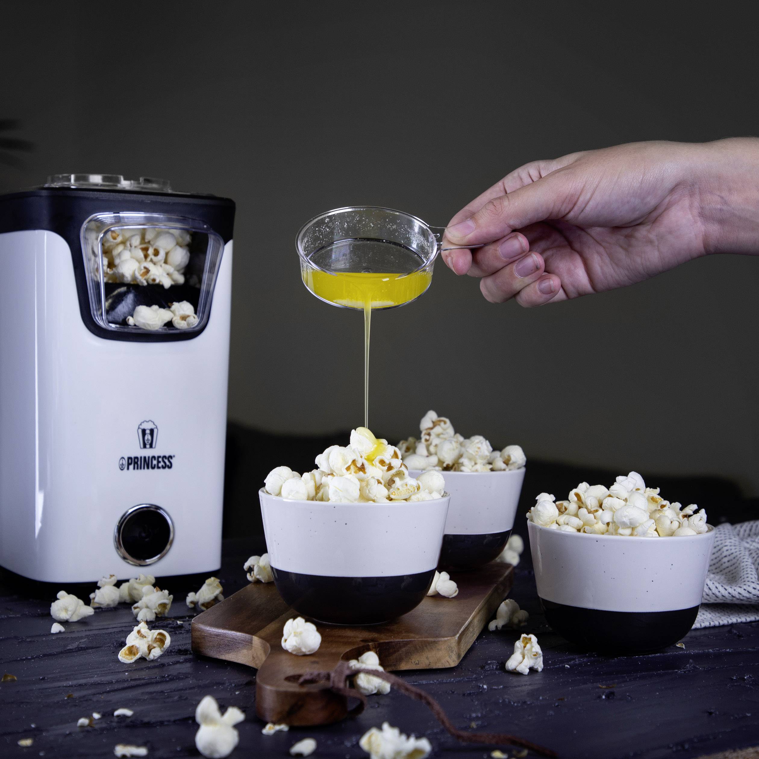 Stroj na popcorn plní misky popcornem. Ruka nalévá roztopené máslo na popcorn. Tmavé pozadí.