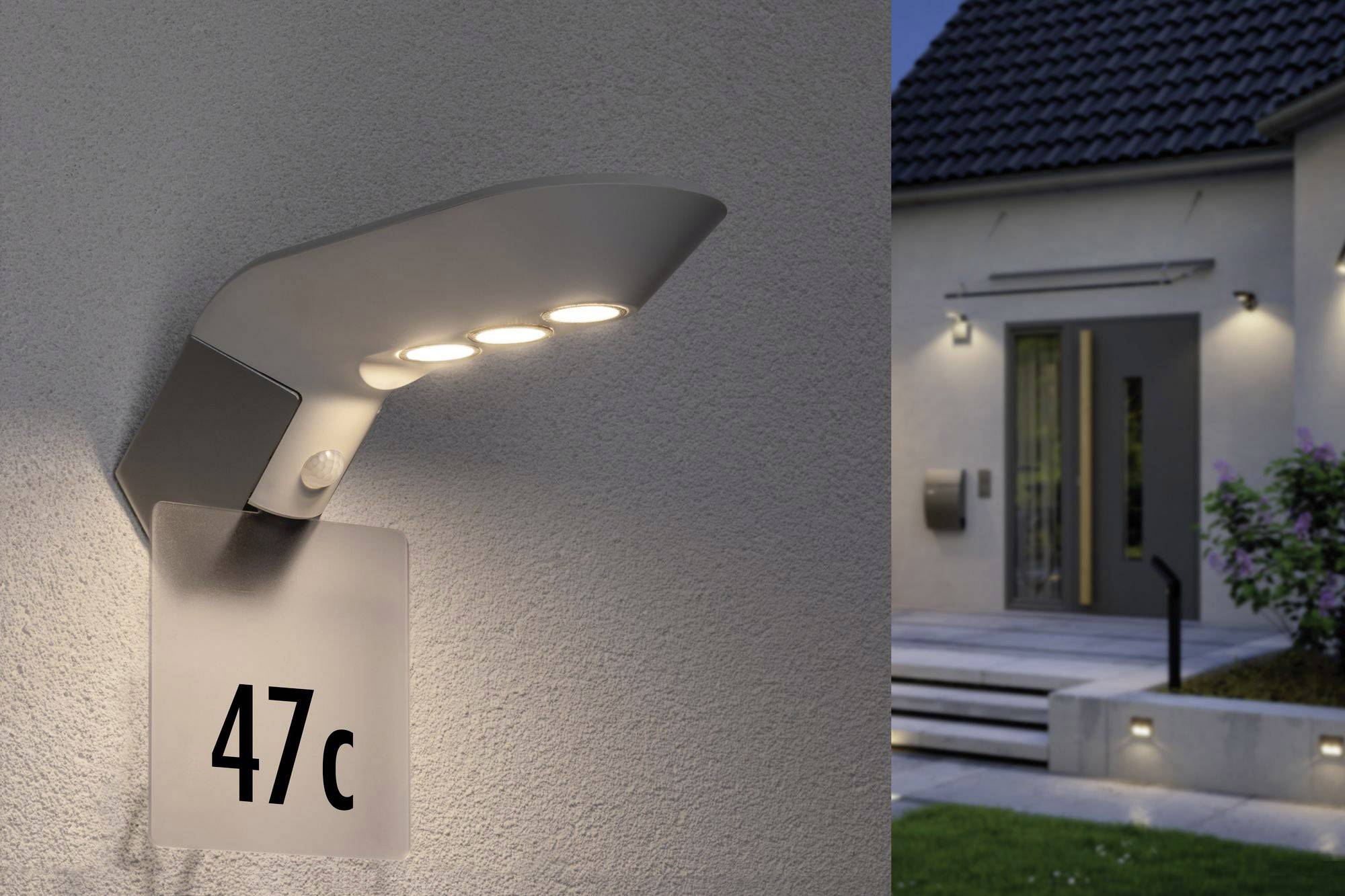 Venkovní lampa s označením '47c' svítí na zdi domu. V pozadí jsou schody vedoucí k hlavnímu vchodu za soumraku.