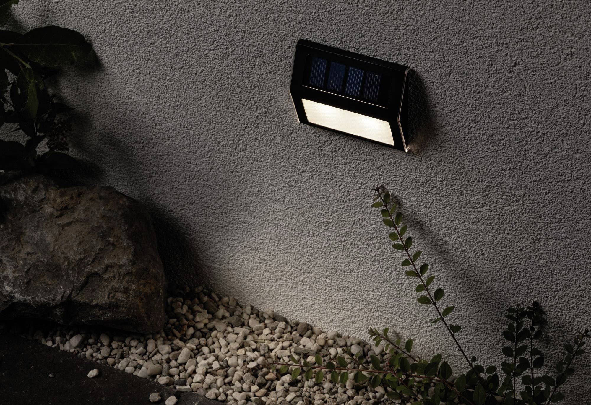 Nástěnná solární lampa osvětluje skalku vedle zdi domu za soumraku. Rostliny a kameny v popředí.