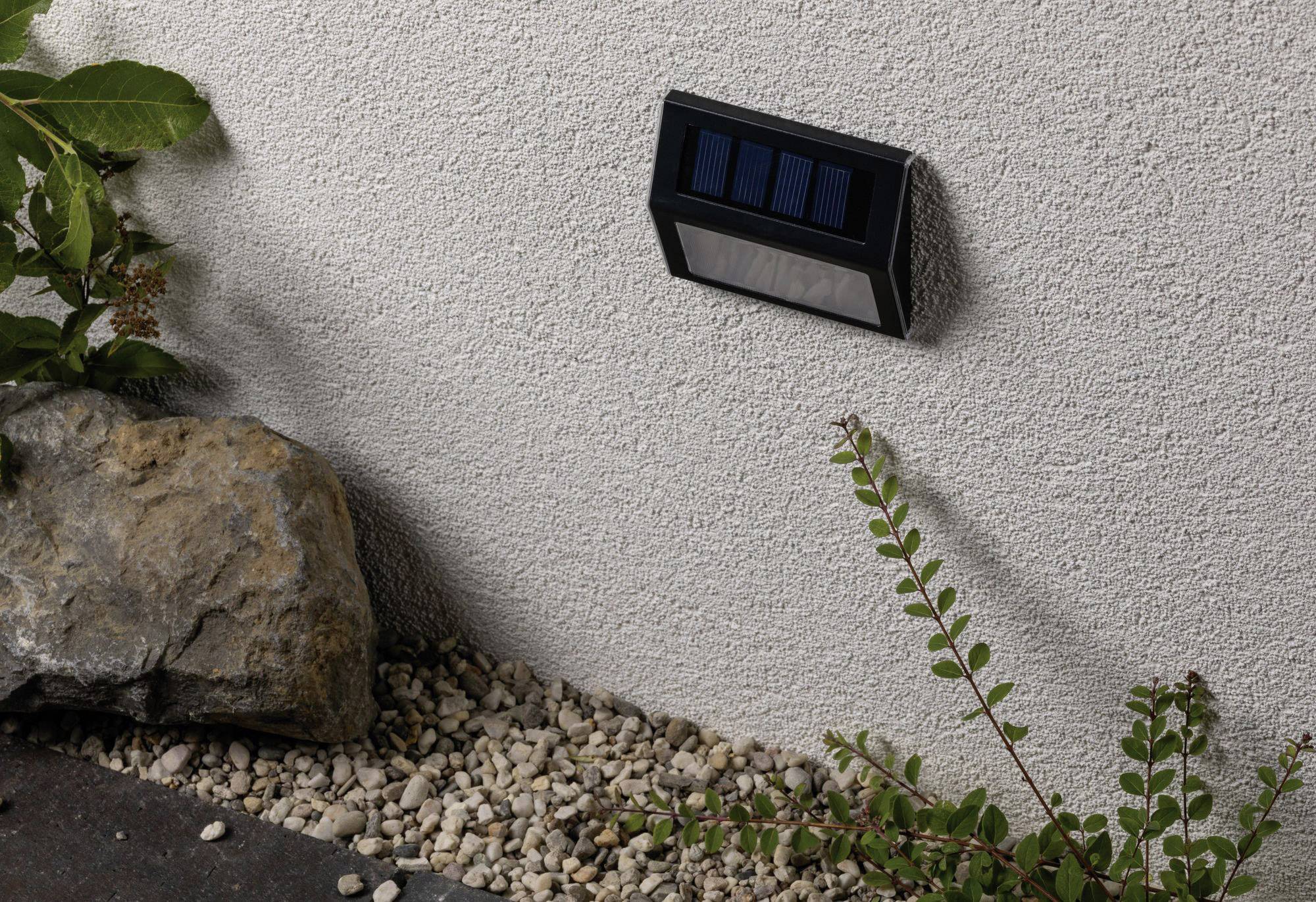 Solární lampa na zdi domu, obklopená kameny a rostlinami. Přes den se dobíjí slunečním světlem a v noci svítí.
