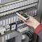 Jedna ruka drží elektrický měřicí přístroj a kontroluje rozvodnou skříň s několika spínači a kabely.