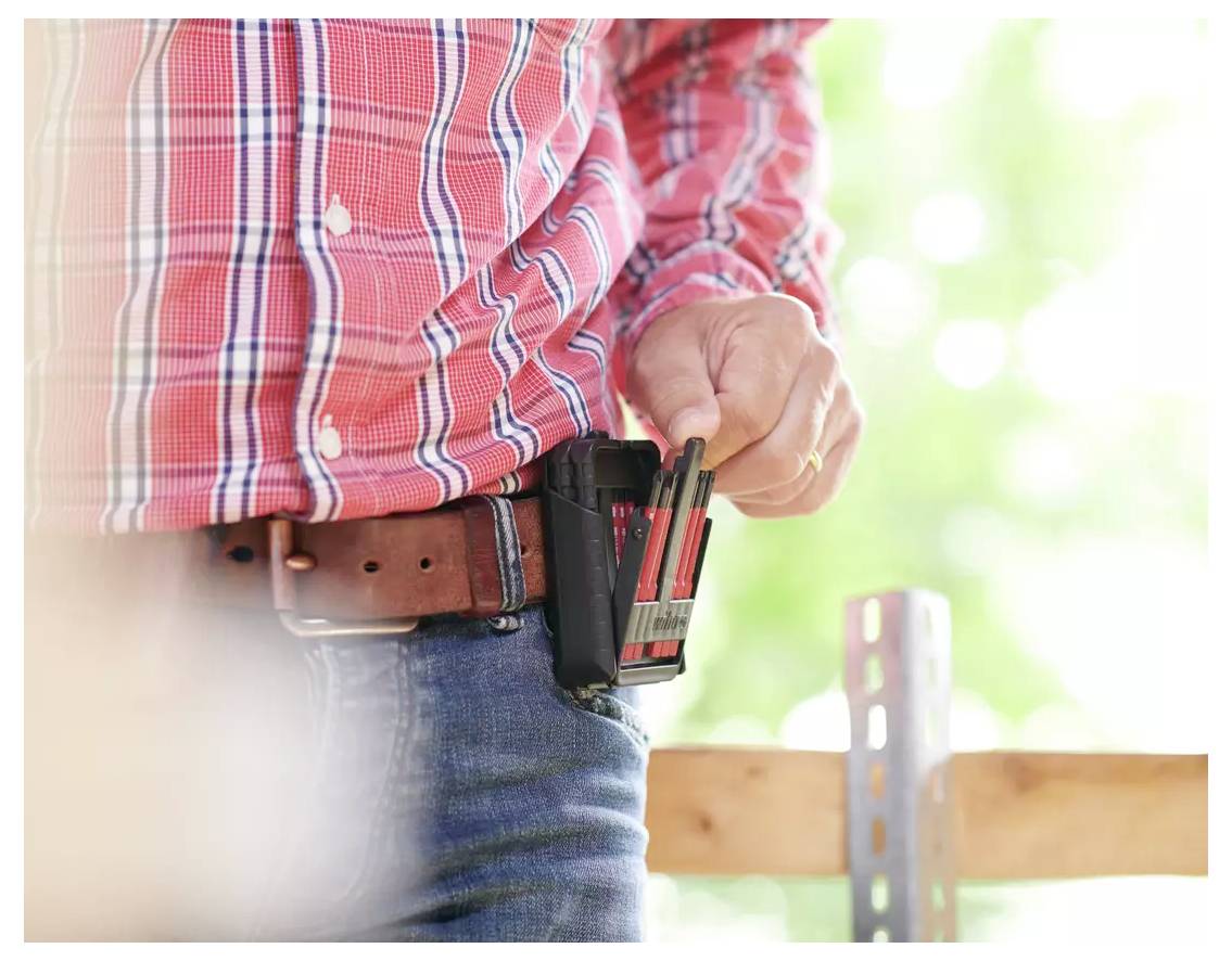 Osoba v kostkované košili a džínech si nese nářadí se tužkami a nástroji na pásu, stojící v jasném venkovním prostředí.