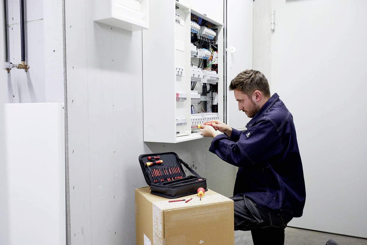 Technik pracuje na otevřené elektrické rozvodné skříni, přičemž jeho kufřík s nářadím stojí na kartonu.