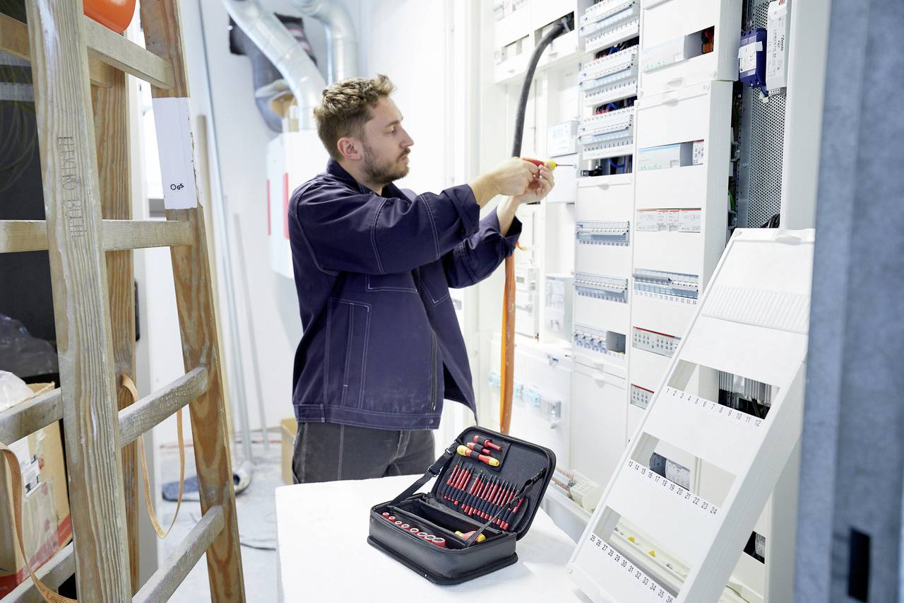 Elektrikář pracuje na rozvaděči, kontroluje kabely a připojení. Otevřený kufřík s nářadím leží na stole před ním.