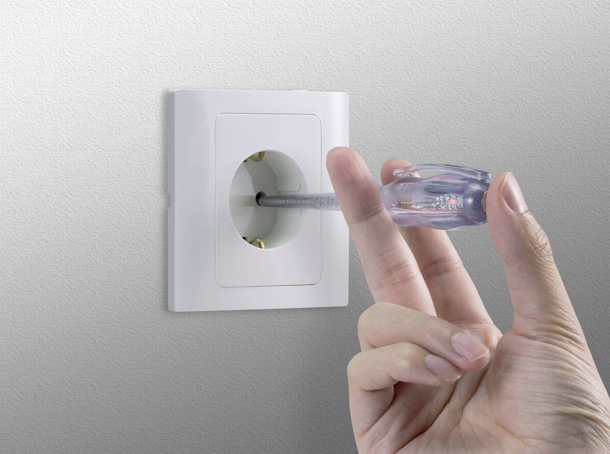Jedna ruka drží zkoušečku napětí zasunutou do elektrické zásuvky ve zdi.