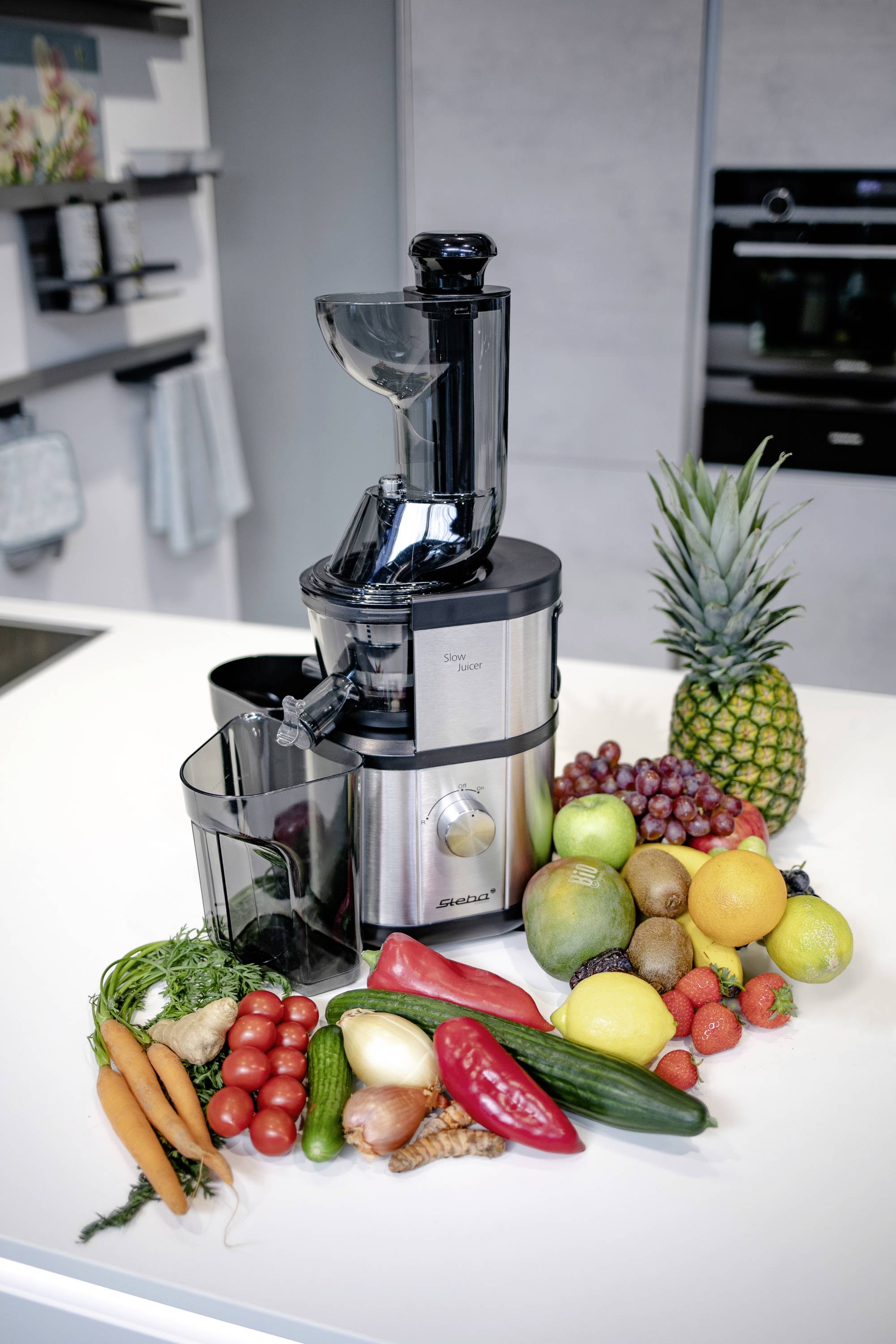 Odšťavňovač na kuchyňské lince, obklopený čerstvým ovocem a zeleninou, jako jsou ananas, hrozny, citróny, mrkev a paprika.