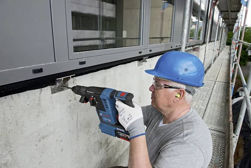 Dělník v modré helmě používá elektrickou vrtačku na betonové zdi mimo budovu, stojící na lešení.