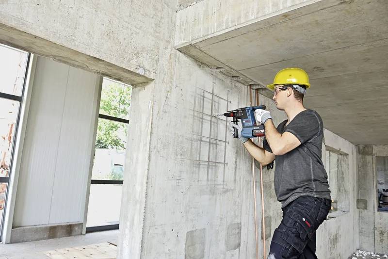Dělník ve stavebním průmyslu s ochrannou přilbou používá elektrickou vrtačku na betonové zdi uvnitř budovy, která je ve výstavbě.