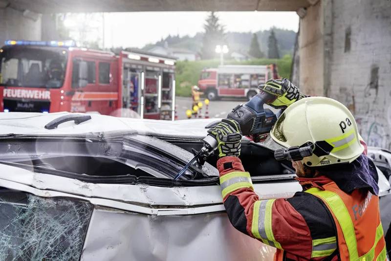 Hasič v ochranném oblečení používá motorové nástroje k řezání boku poškozené vozidla. V pozadí jsou viditelné hasičské vozy.