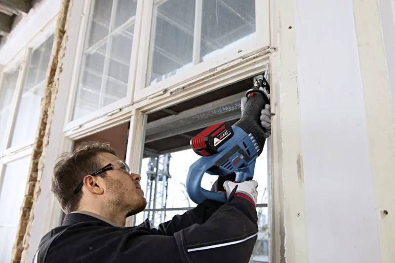 Osoba používající elektrický nástroj při práci na okenním rámu, oblečená v ochranných brýlích a rukavicích, soustředěná na renovaci v konstrukčním prostředí.