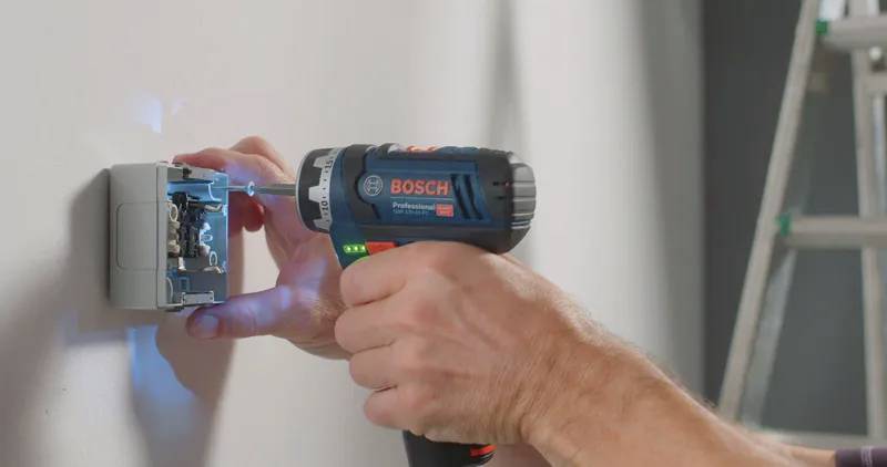 Osoba používá elektrickou vrtačku Bosch k instalaci nebo opravě elektrické zásuvky na stěně, v pozadí je vidět žebřík.