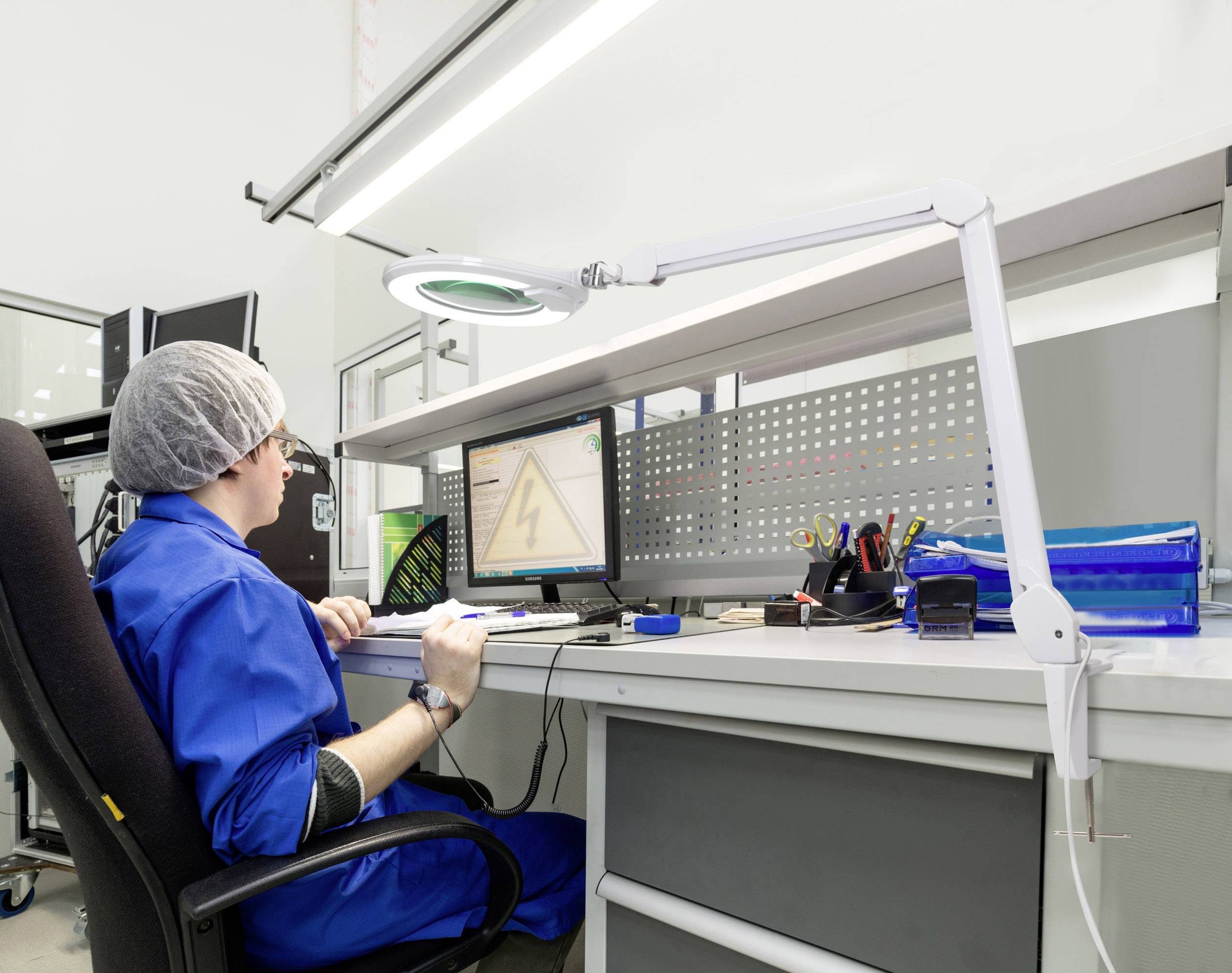 Osoba v modré ochranné kombinéze a s vlasovou síťkou pracuje na počítači v laboratorním prostředí. Na pracovním stole jsou nástroje a monitor.