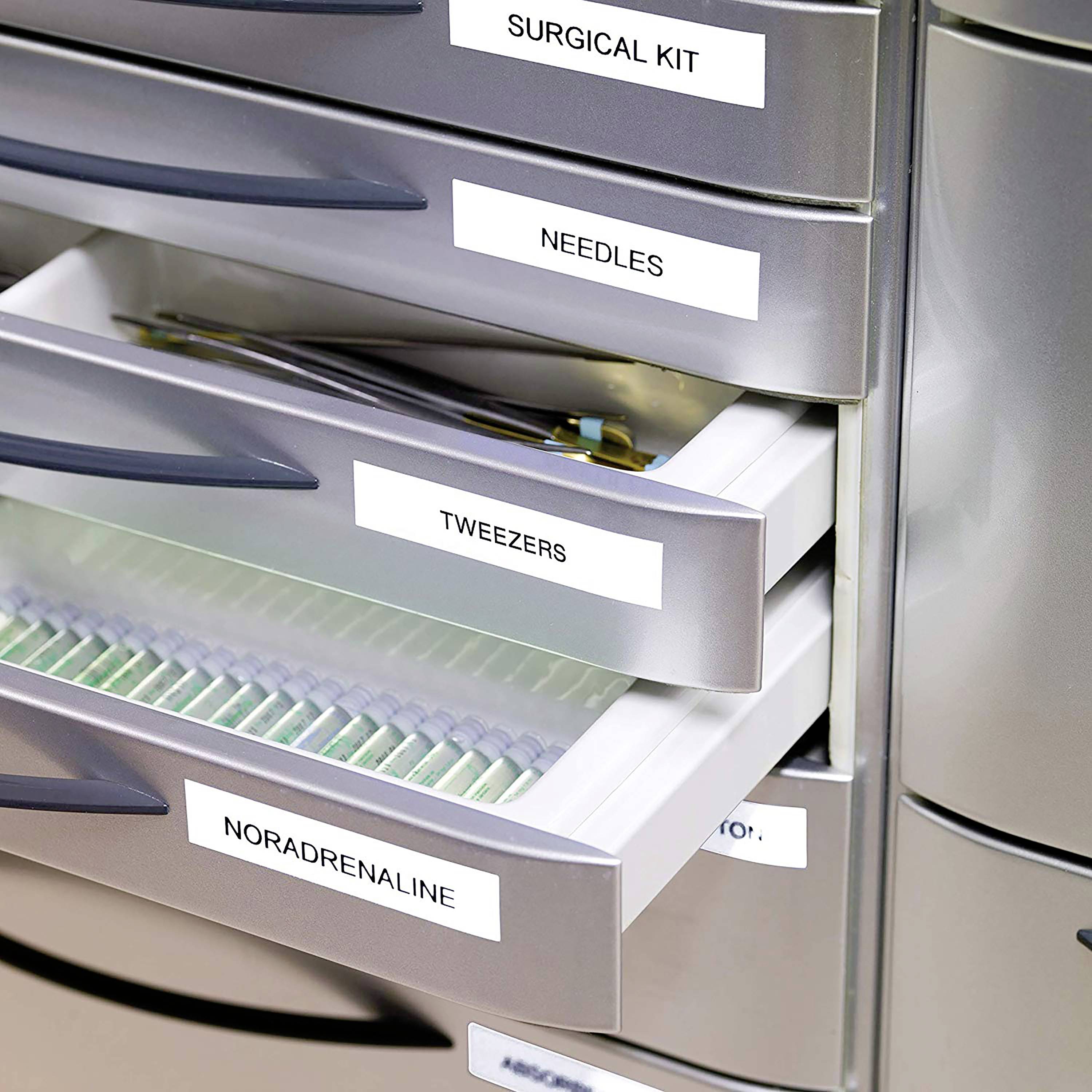 Lékařská skříňka s několika zásuvkami, označenými štítky 'Chirurgická sada', 'Jehly', 'Pinzety', 'Noradrenalin'. Některé zásuvky jsou pootevřené.