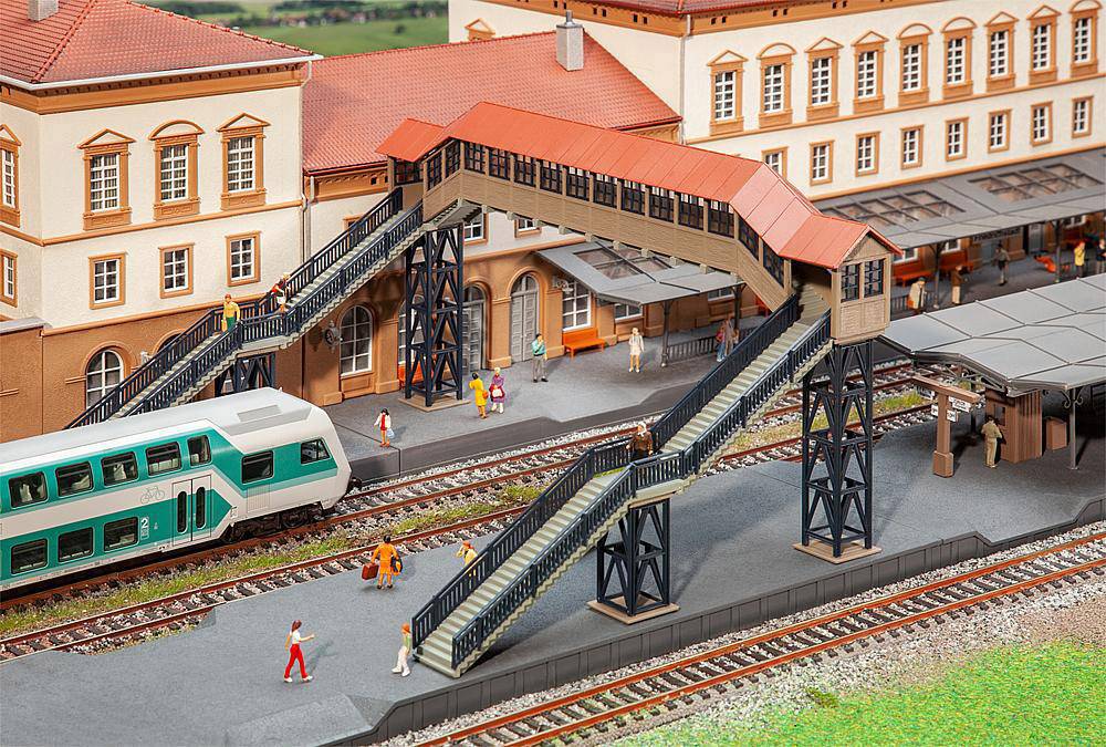 Model kolejové železnice zobrazuje nádraží s vlakem, lávkou pro pěší a figurkami. Hlavní budova má červené střechy. Lidé se pohybují na lávce a kolejích.
