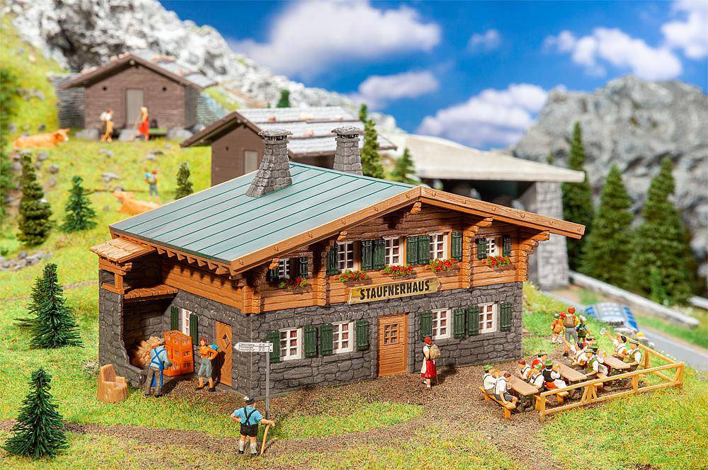 Detailní miniaturní model ukazuje horskou chatu s nápisem 'Staufenhaus'. Lidé sedí na lavičkách, zatímco ostatní prozkoumávají okolí. V pozadí jsou vidět hory a stromy.