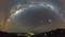 Panoramatický snímek noční oblohy ukazuje Mléčnou dráhu jako zářící oblouk nad krajinou s třpytícími se městskými světly.