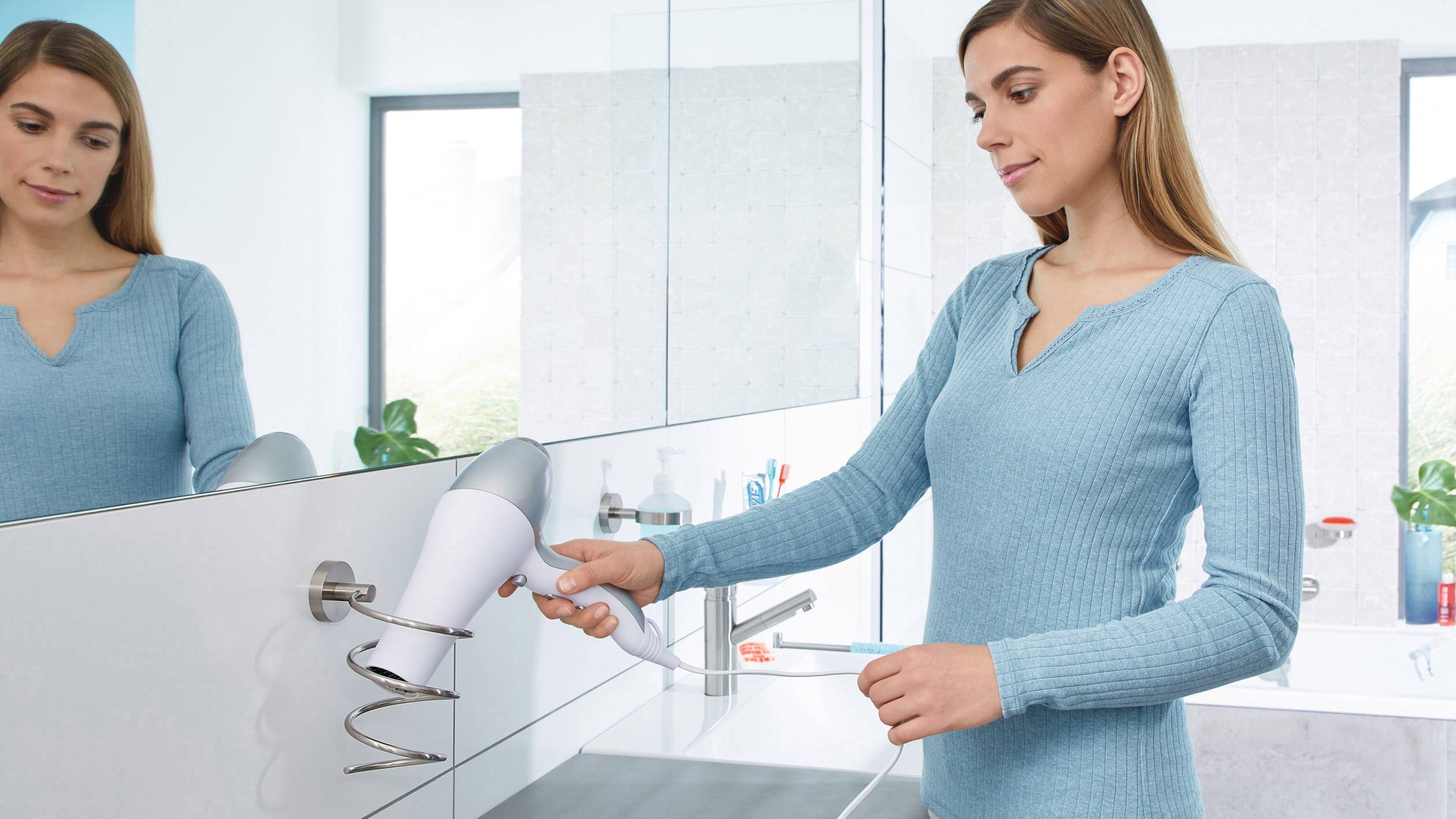 Žena v modrém svetru upevňuje fén do nástěnného držáku v koupelně.
