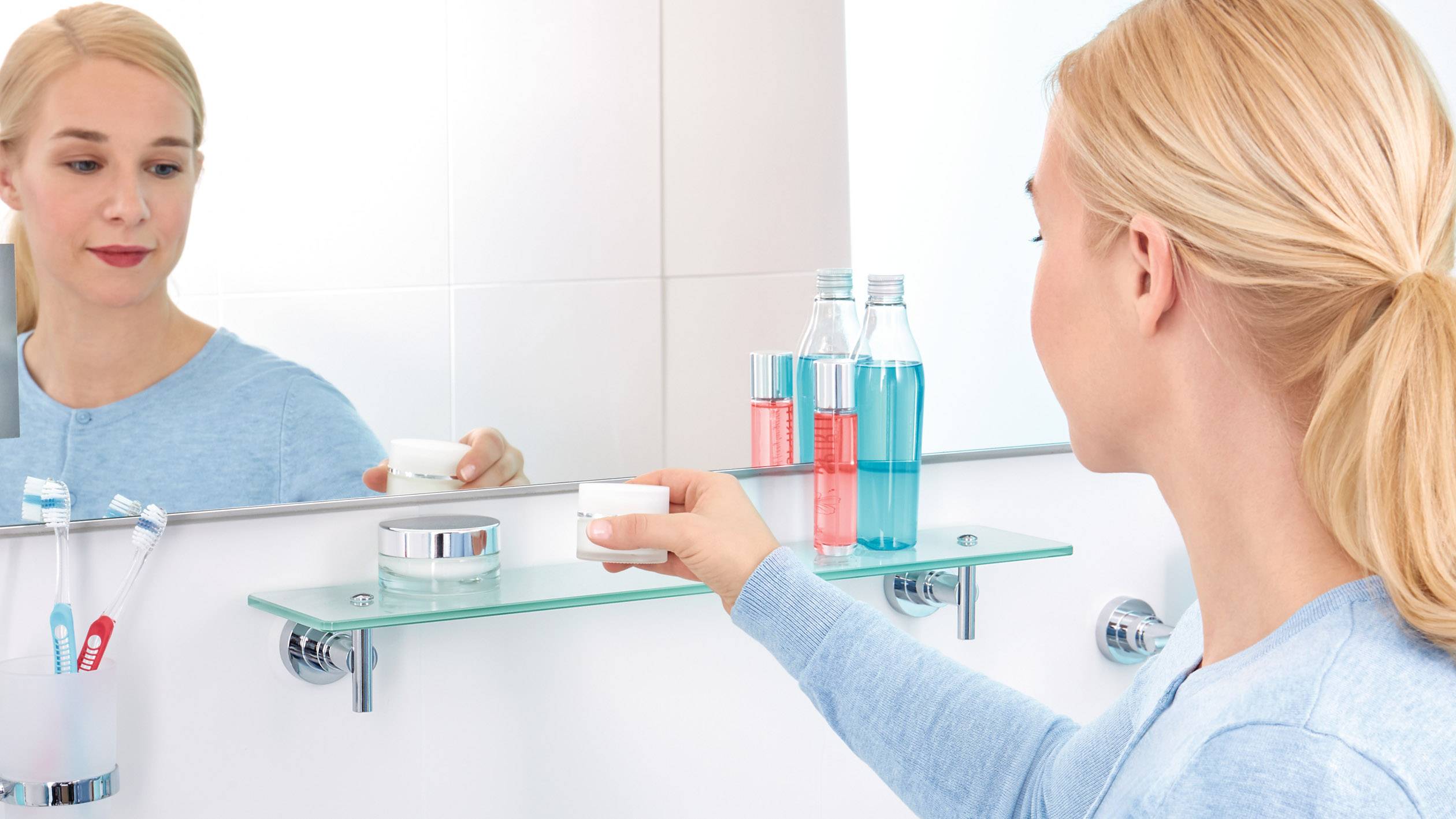 Žena v koupelně sundává dózu s krémem ze skleněné police. Vedle stojí zubní kartáčky a ústní voda.