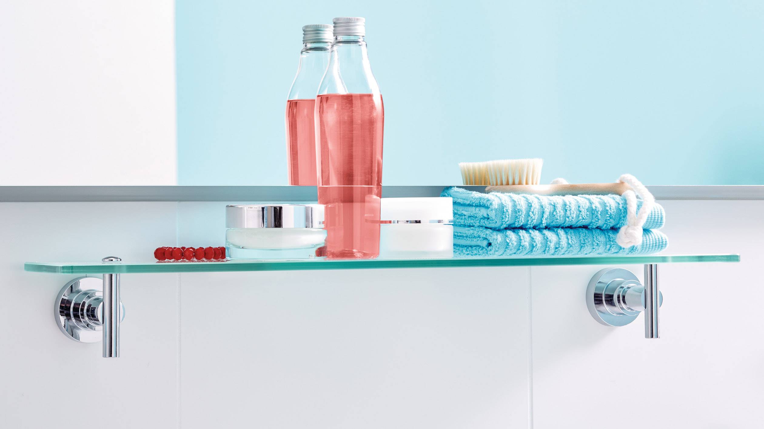 Hygienické výrobky, včetně dvou růžových lahví, kelímku, koupací osušky a kartáče, jsou umístěny na skleněné polici v koupelně.